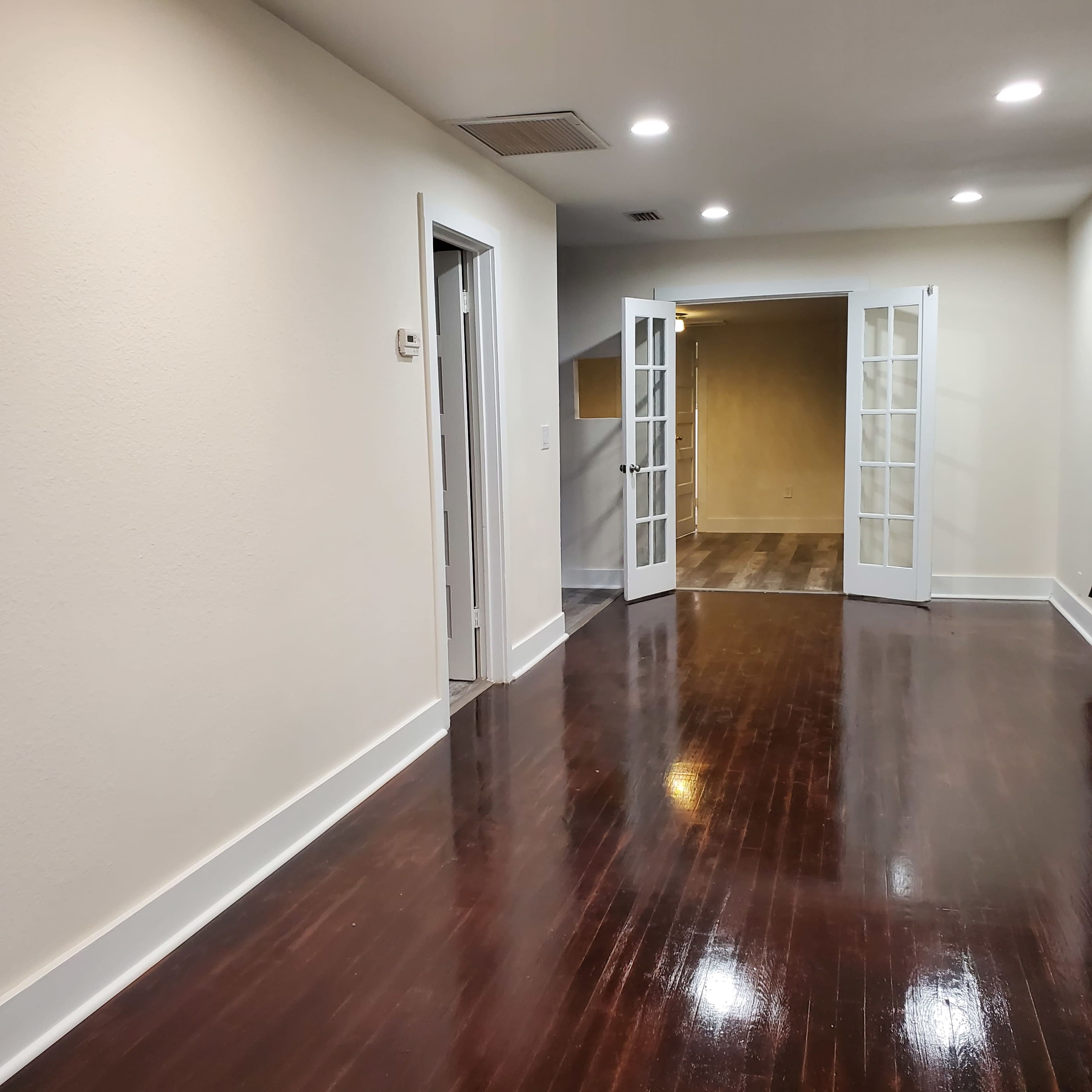 A well-lit hallway with polished wooden floors leads to a room with double French doors.