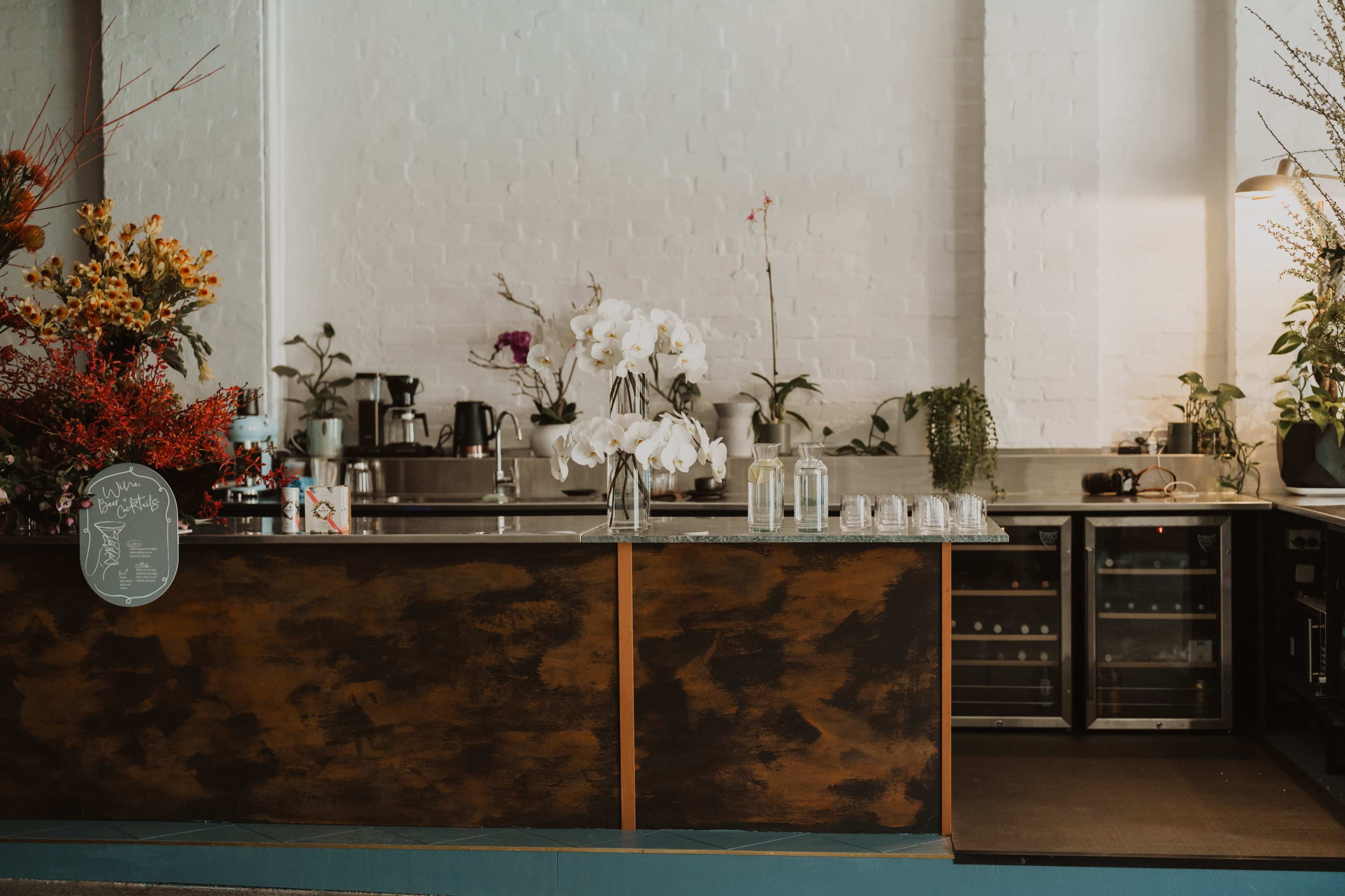 A coffee shop counter with floral arrangements and various kitchen appliances visible in a minimalist setting.