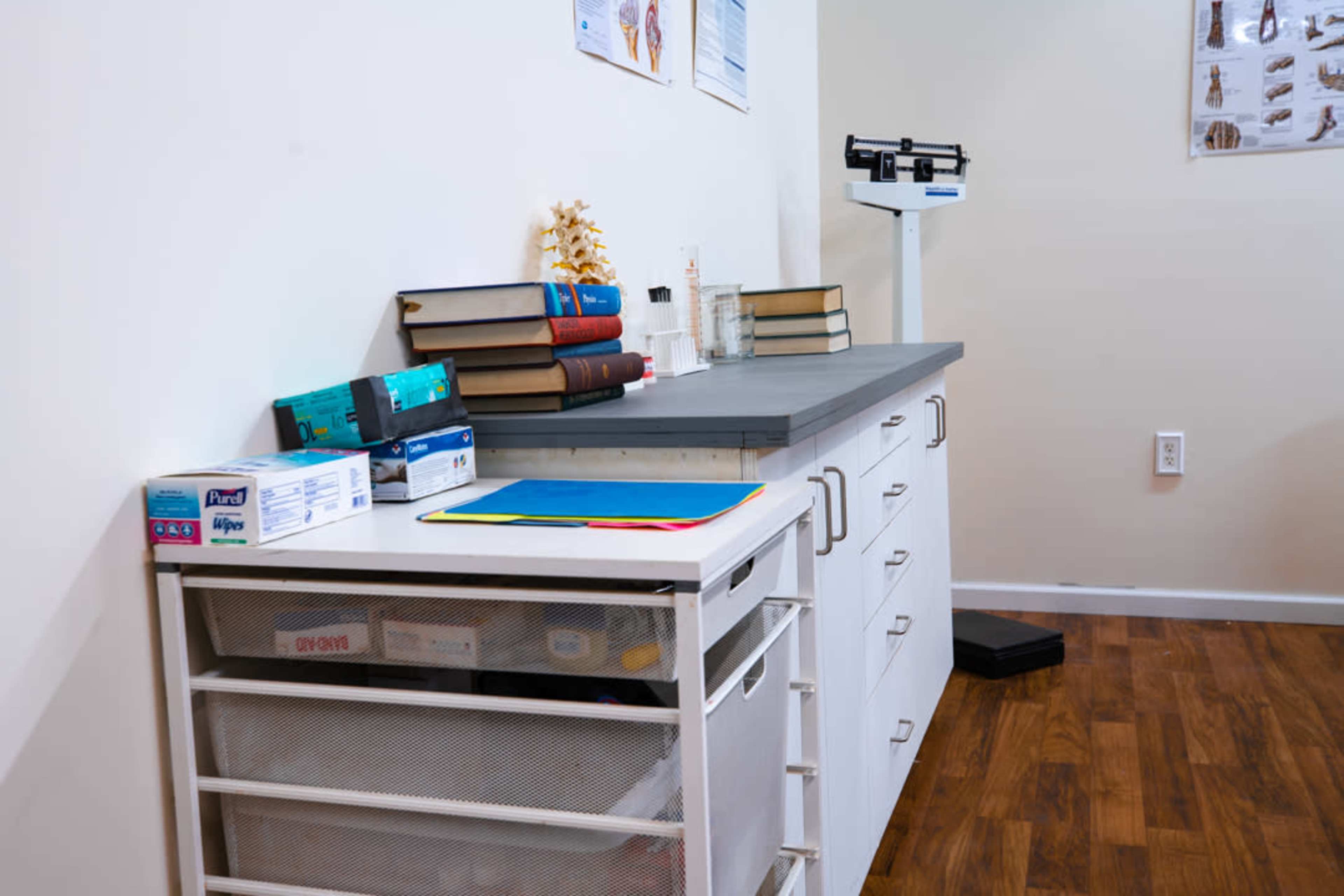 A medical examination room features a countertop with stacked books, a spine model, and medical supplies, alongside a scale and storage drawers.