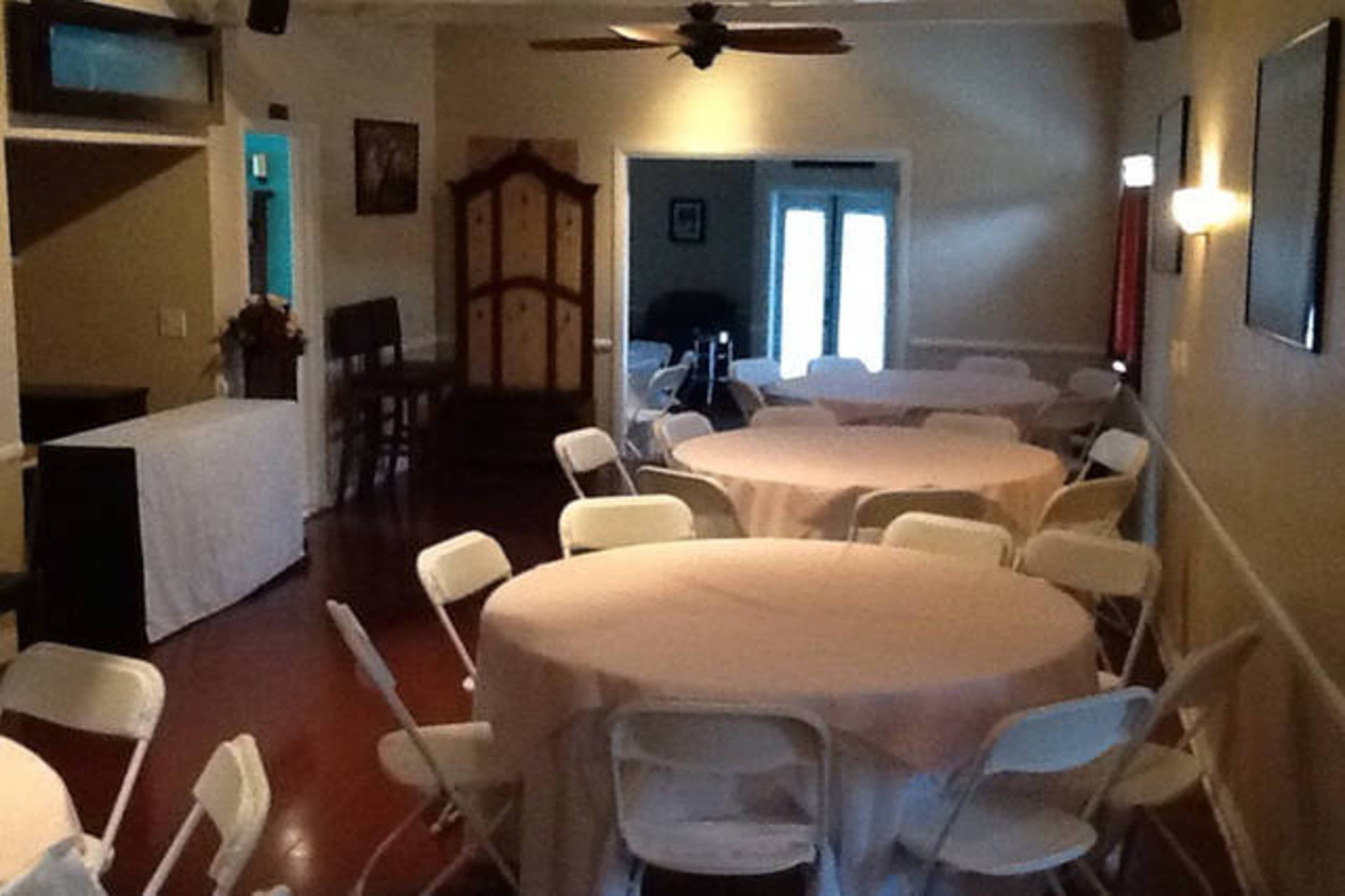 A spacious room features several round tables covered with cloths and white folding chairs arranged around them, with a bar area and an entrance in the background.