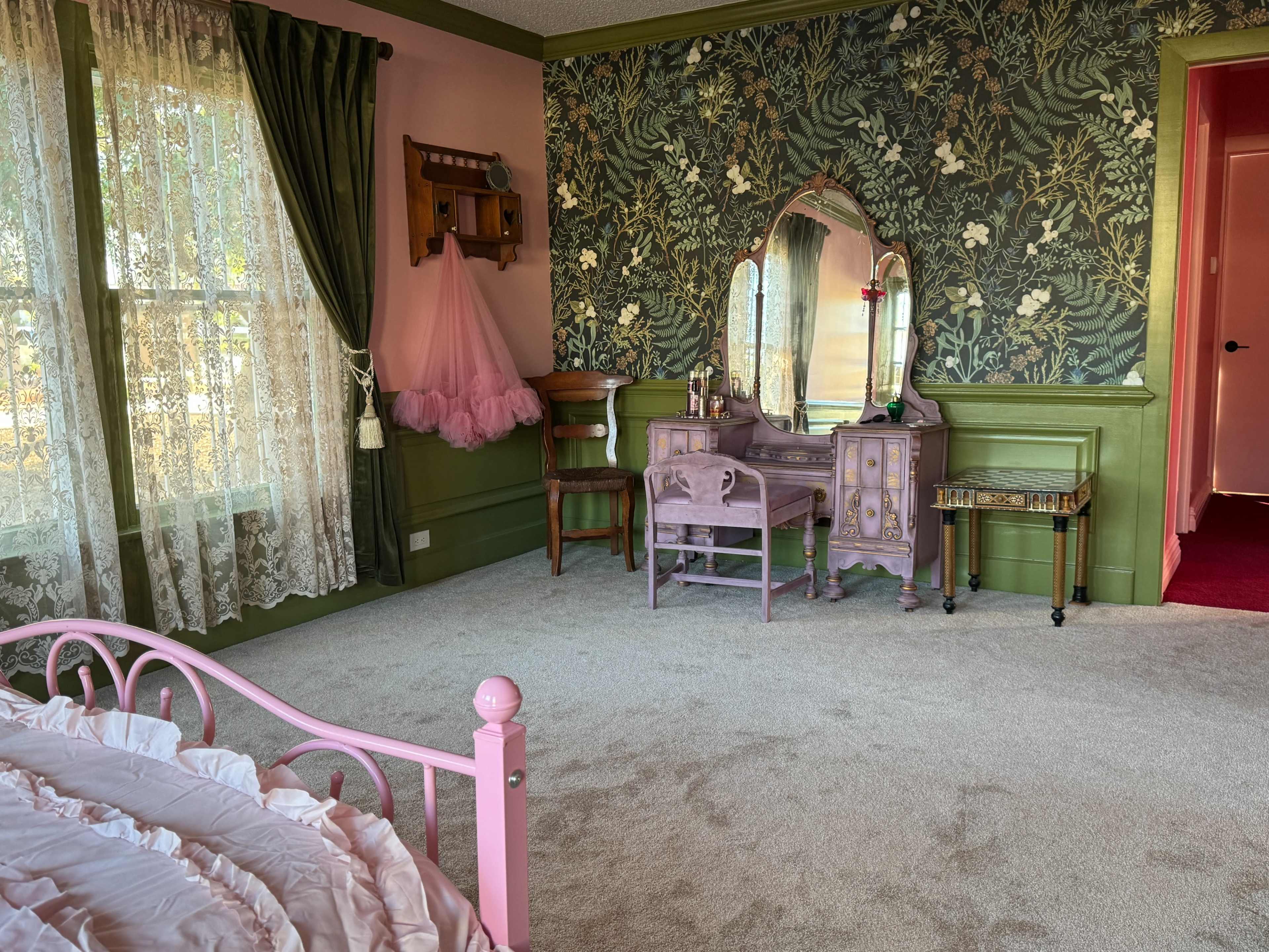 The room features pink walls, a floral wallpaper accent, a vintage vanity with a mirror, and soft beige carpeting.