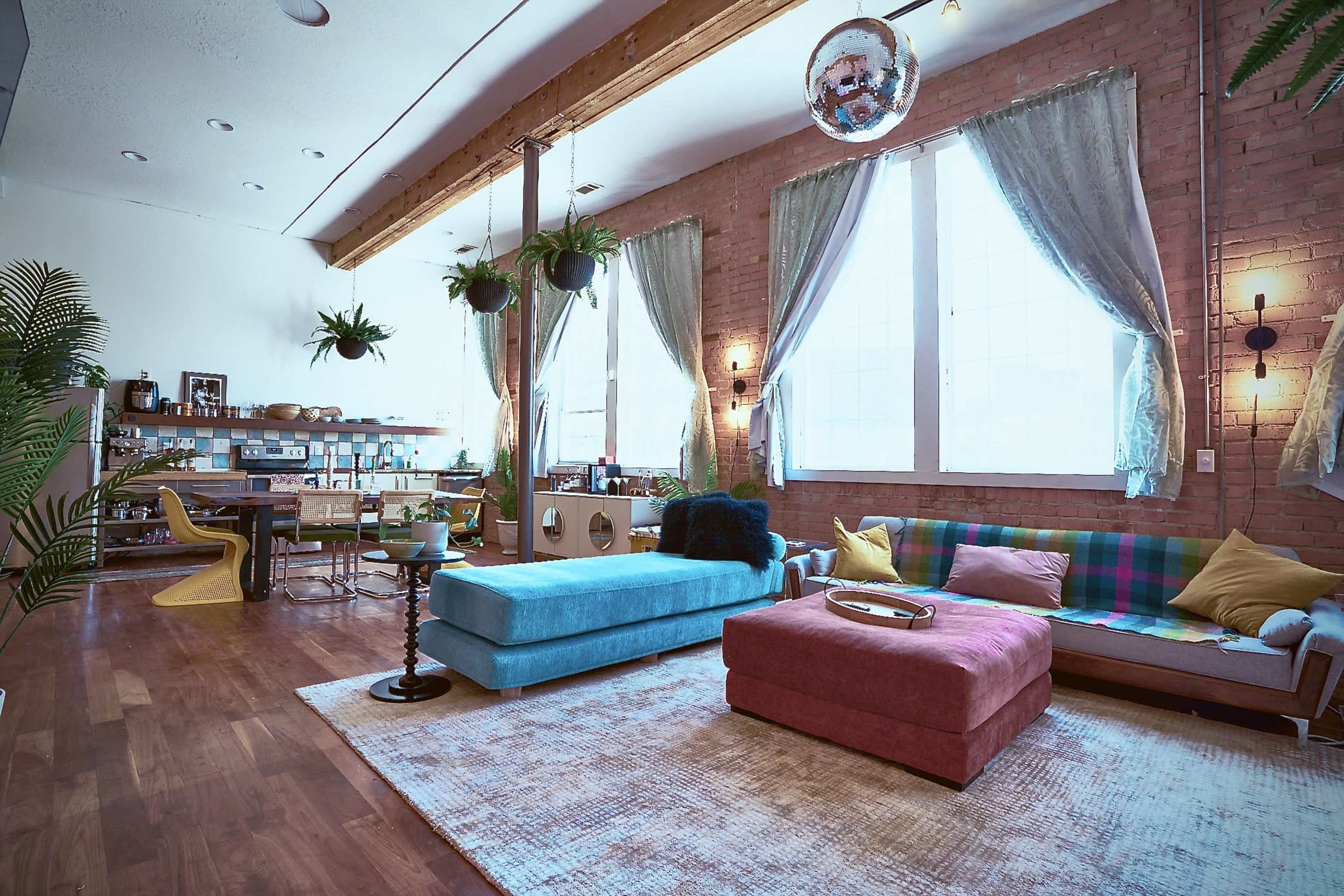 A modern living room with a blue sofa, a pink ottoman, large windows, and various plants hanging from the ceiling.