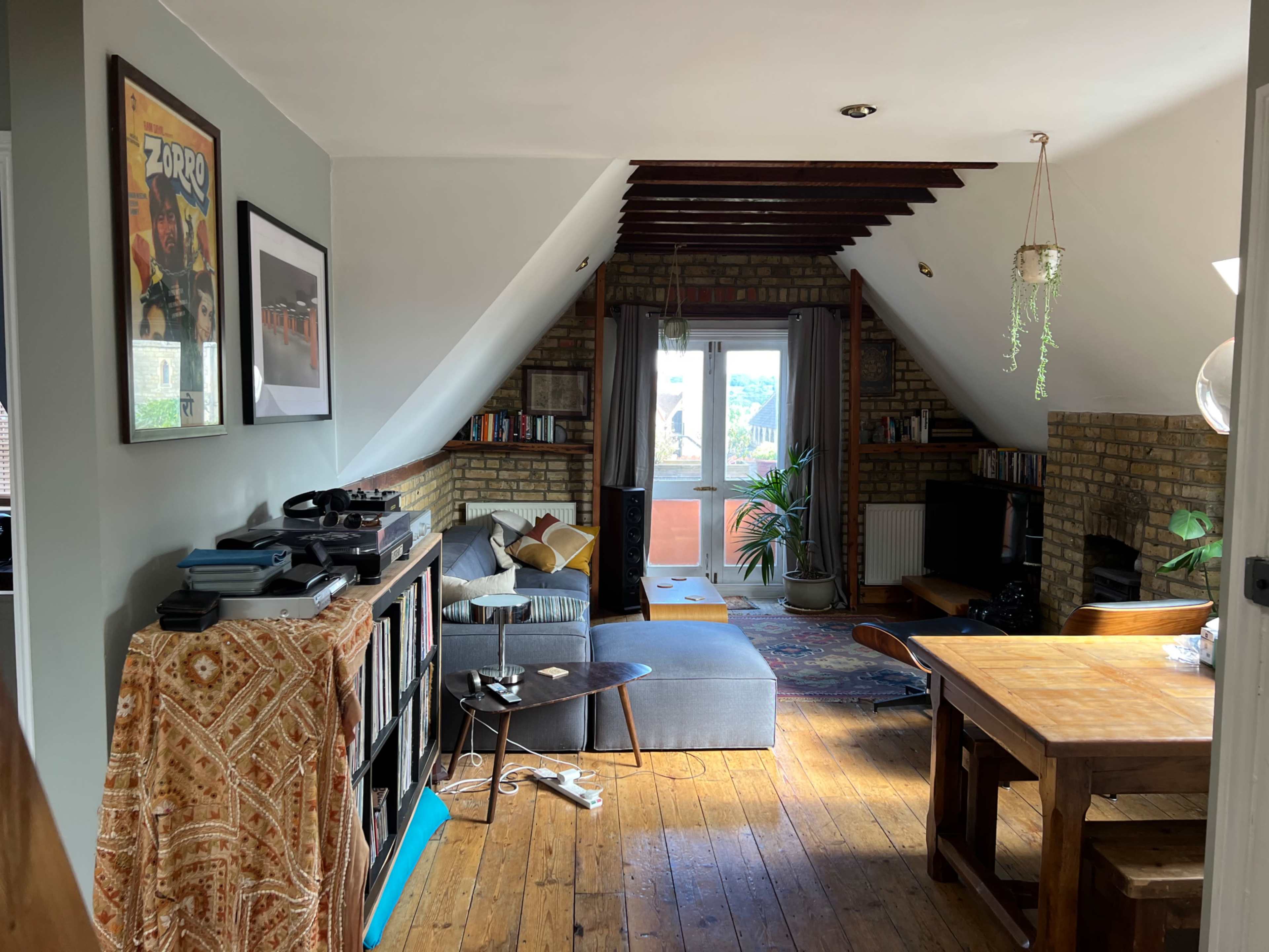 The image shows an attic living space with wooden floors, a seating area, a dining table, and shelves filled with books and decor.