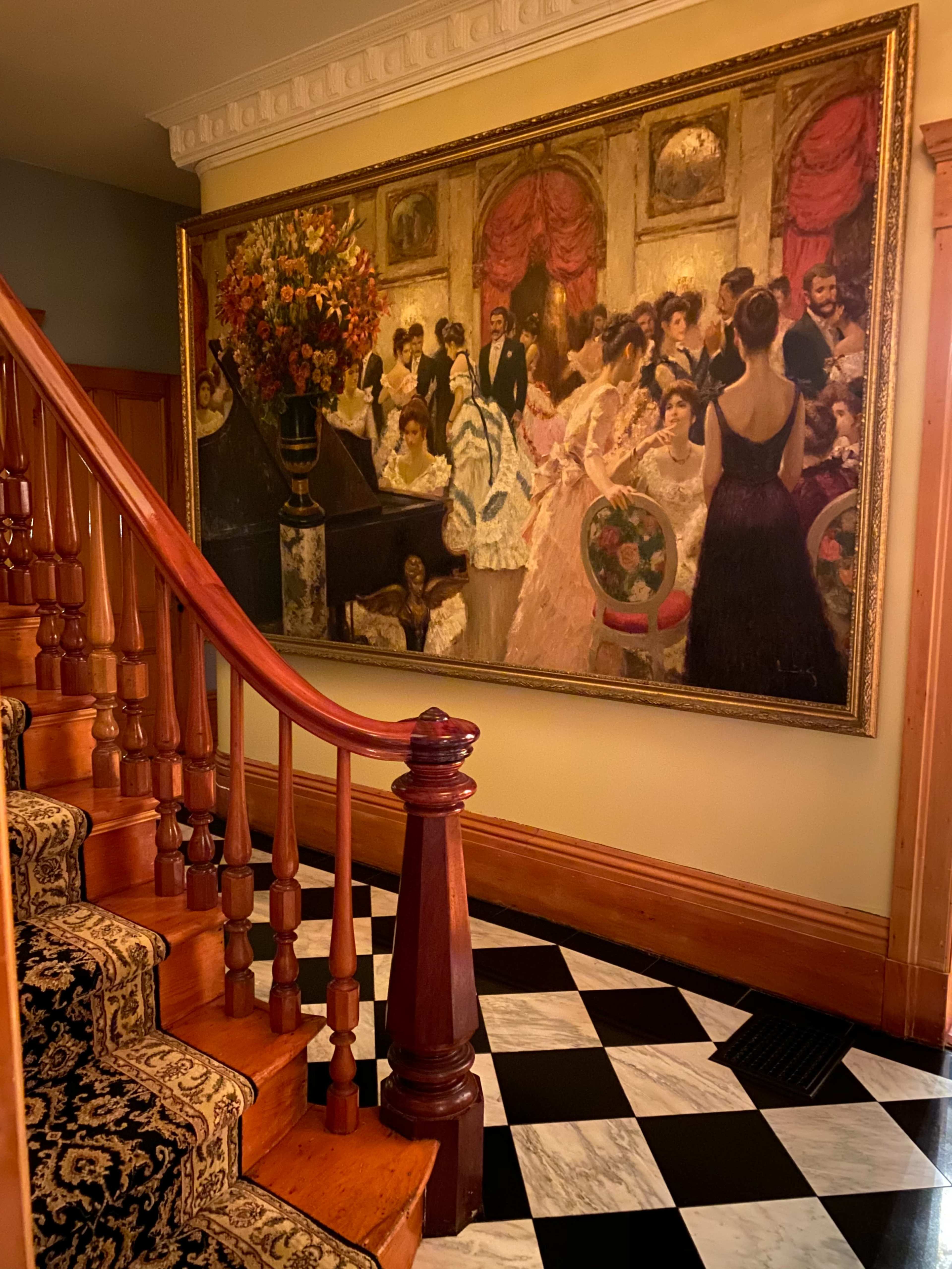 A large painting depicting a formal gathering hangs on the wall above a wooden staircase with a patterned runner, set against a checkered black and white floor.