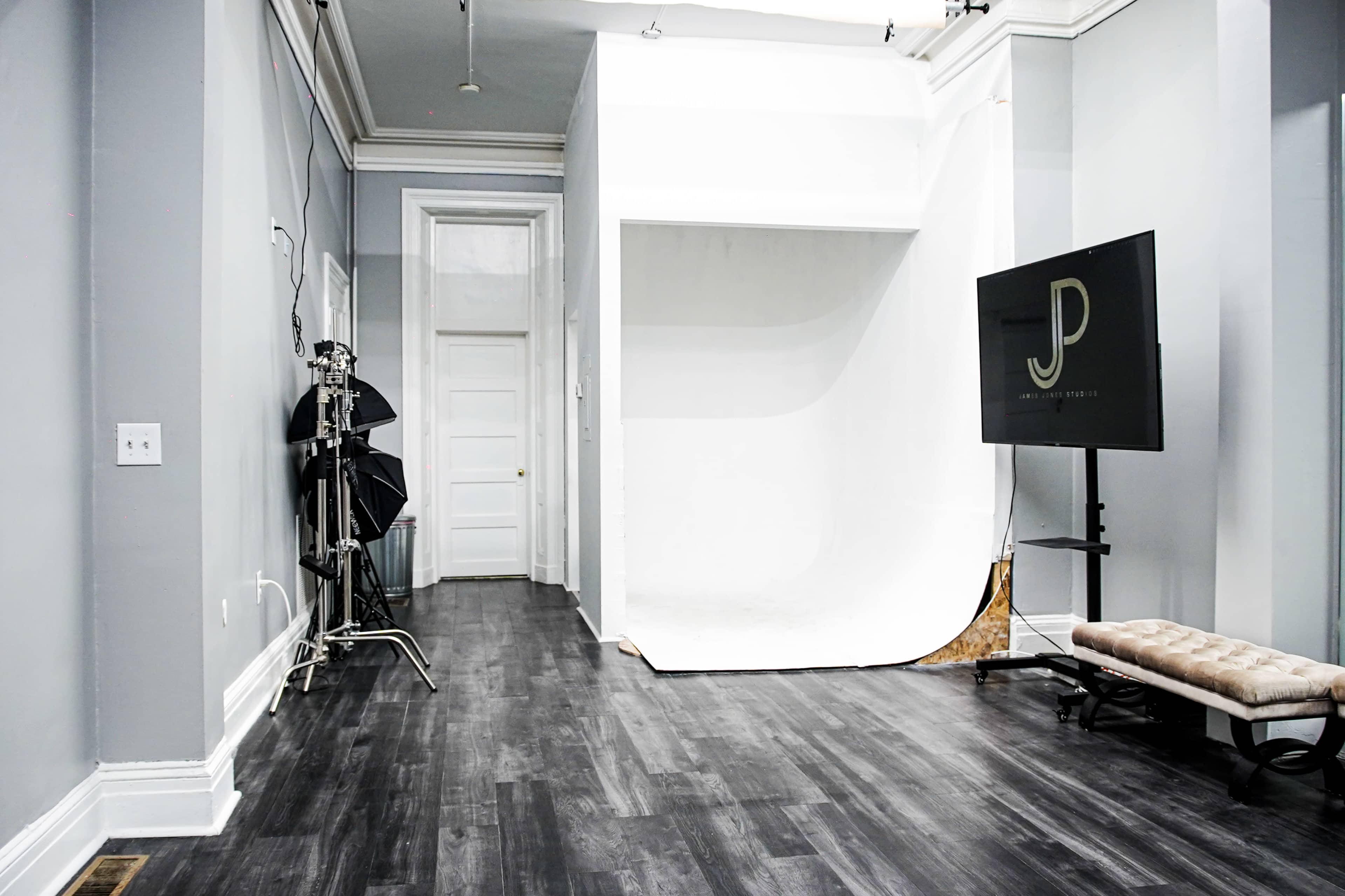 A photography studio with a white backdrop, a black lighting equipment stand, and a television displaying a logo, all set in a room with gray walls and dark wood flooring.