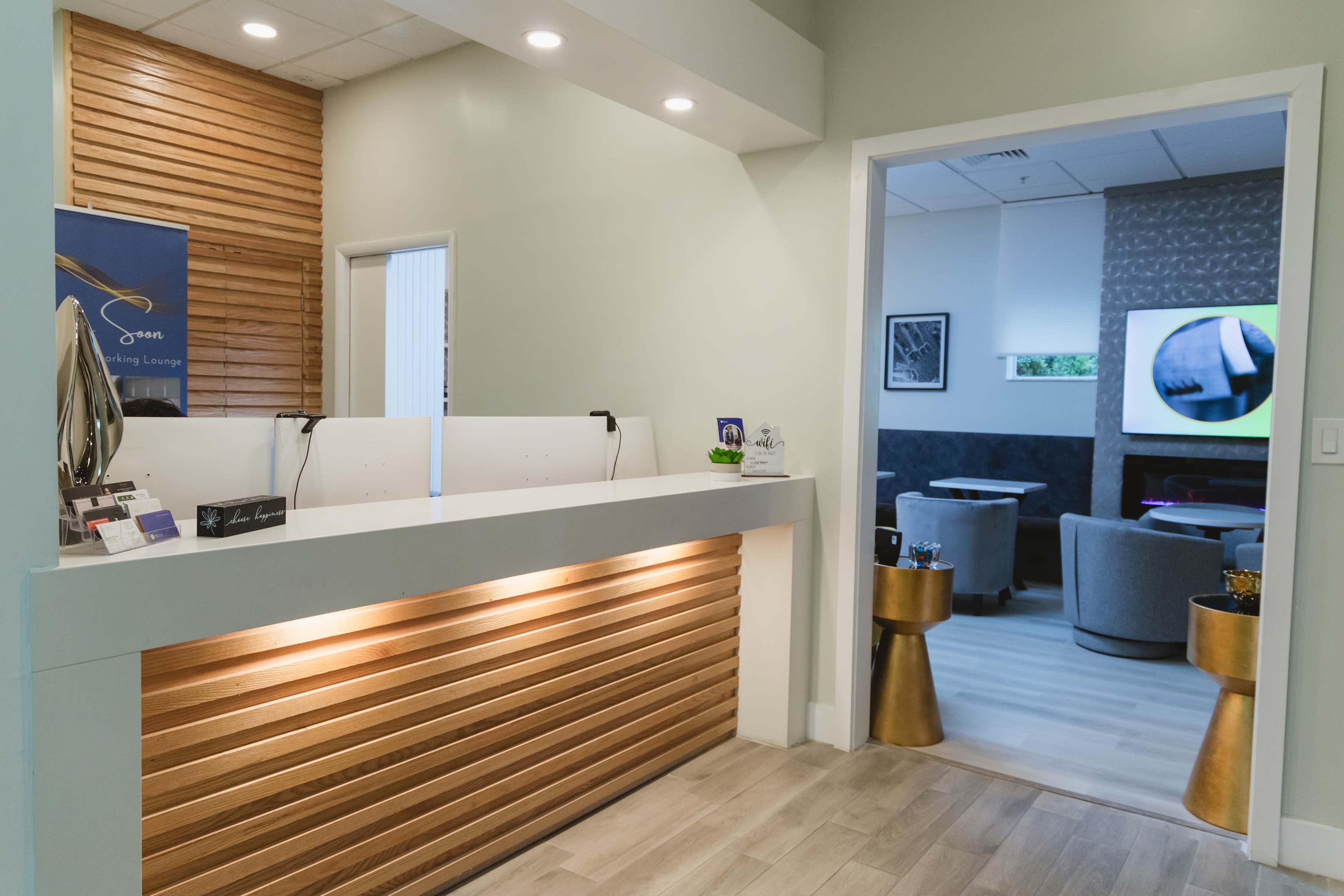 The image shows a modern reception area with a wooden front desk and a view into a lounge with comfortable seating and a television.