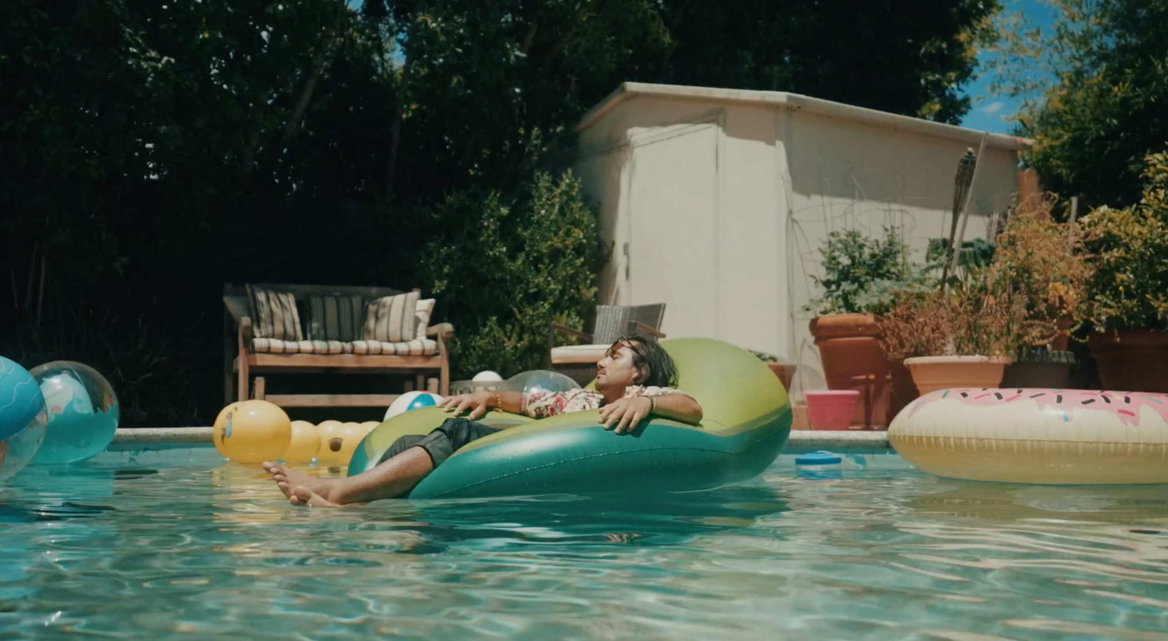 A person relaxes on a green inflatable float in a swimming pool surrounded by colorful pool toys and a shaded seating area.