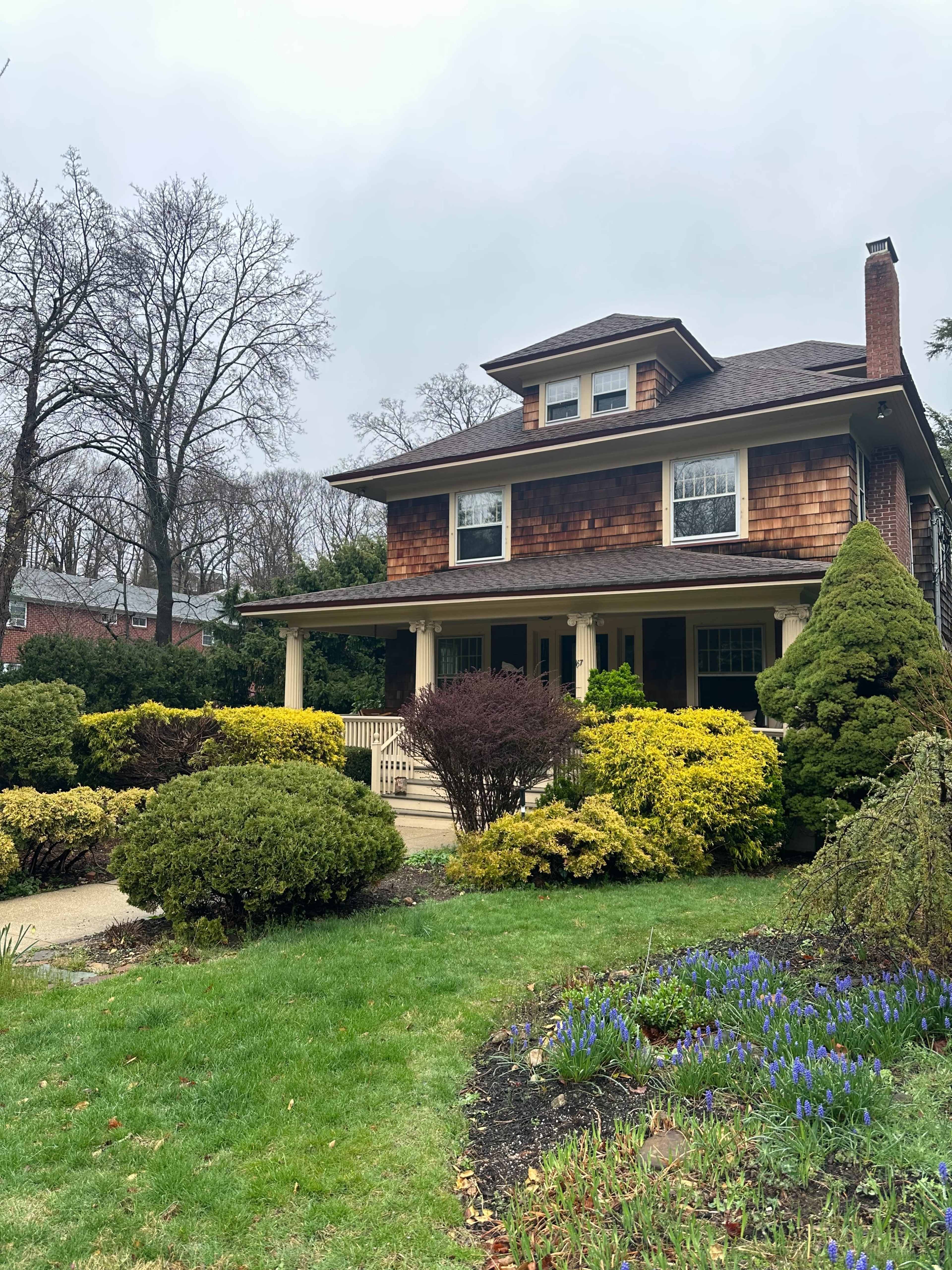 The image shows a two-story house with a brown shingle exterior, surrounded by well-maintained shrubs and colorful flowerbeds in a residential neighborhood.