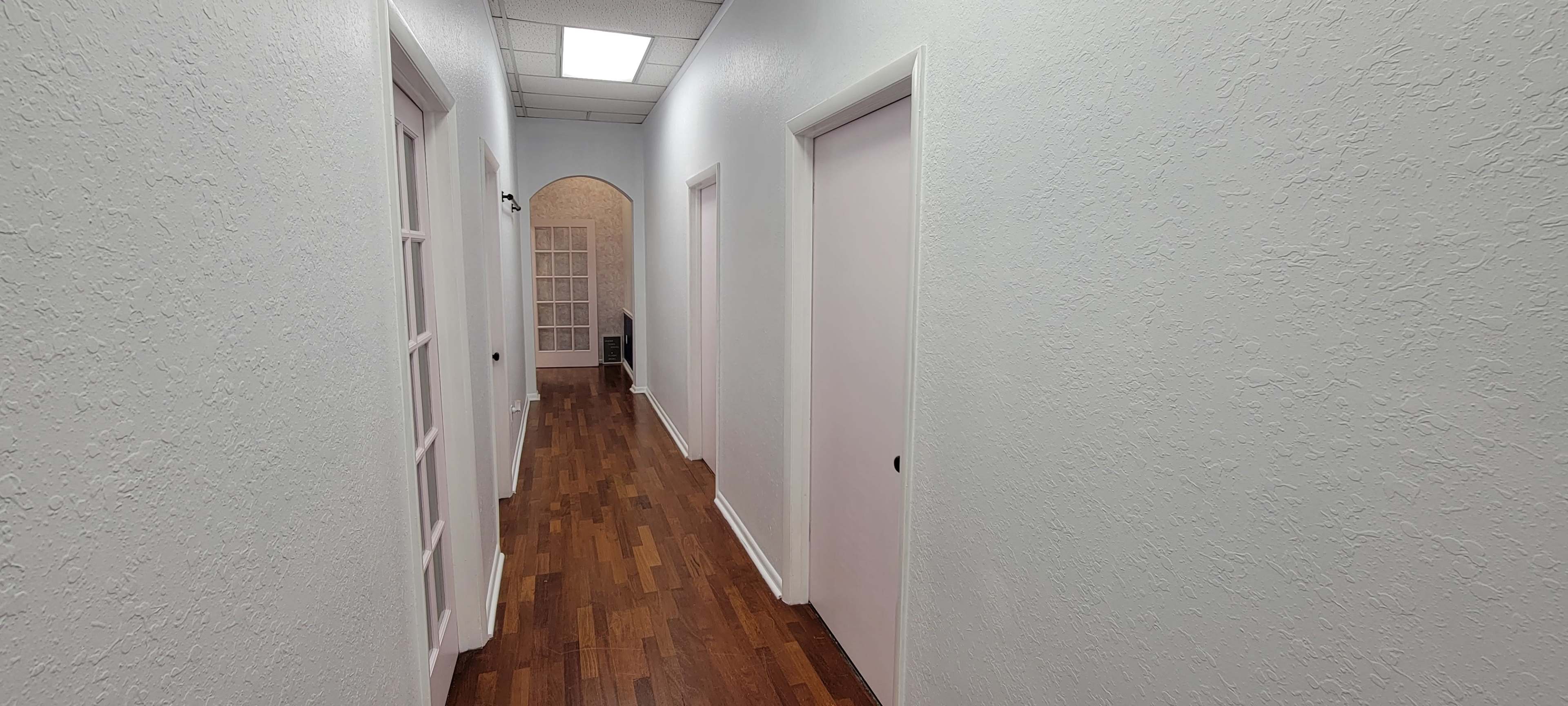 A well-lit corridor features white walls, wooden flooring, and doors on either side leading to different rooms.