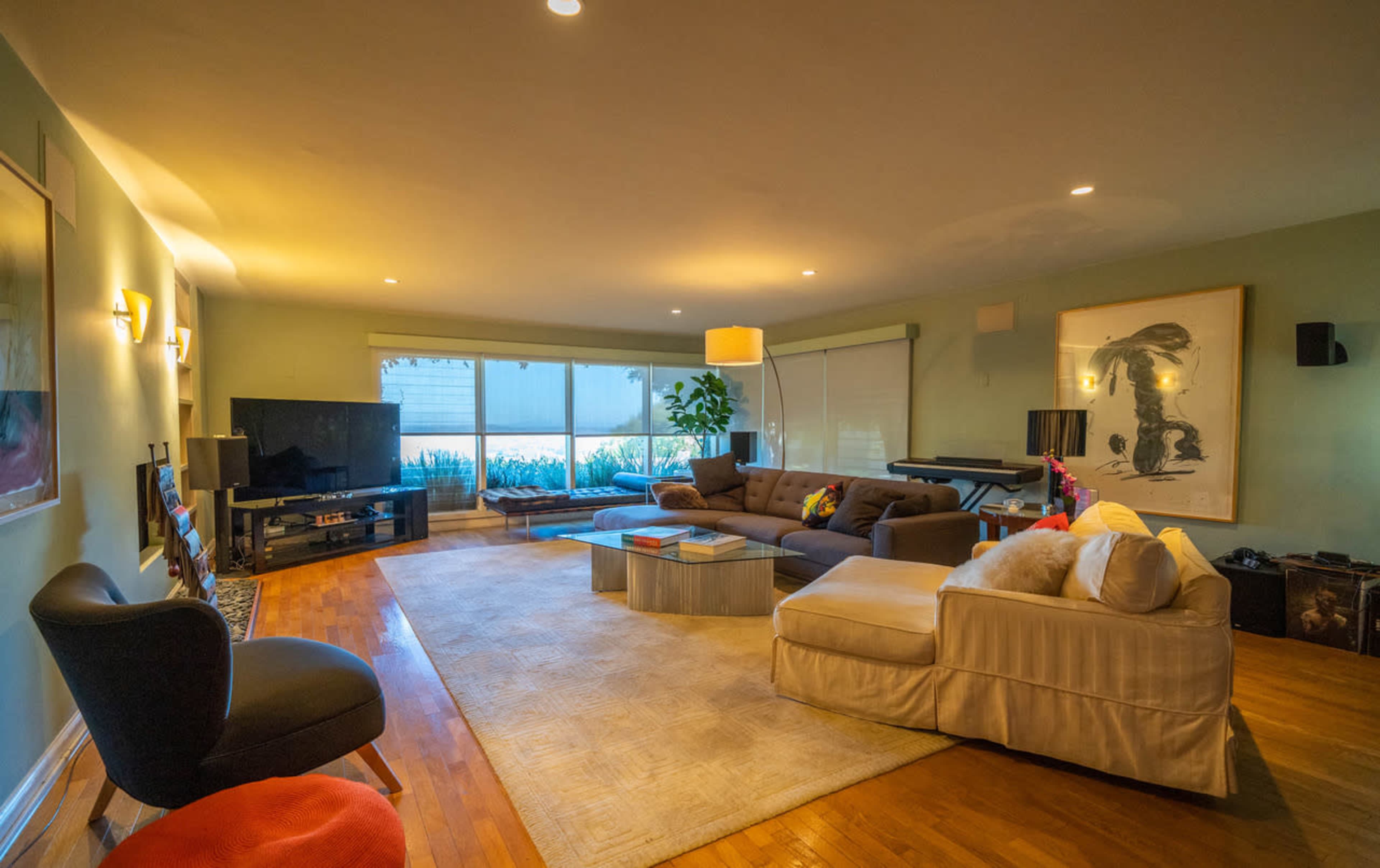 The room features a spacious living area with a sectional sofa, a large television, and windows allowing natural light, complemented by a green wall and artwork.