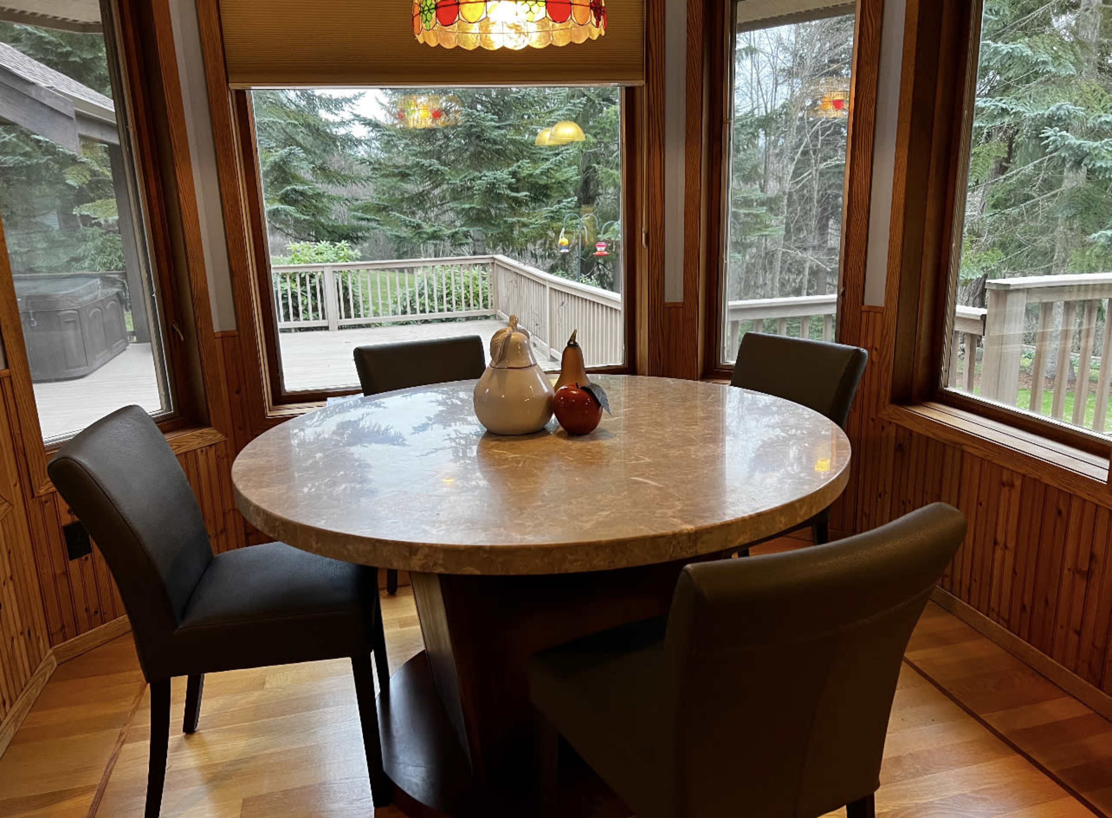 The scene features a round marble dining table surrounded by four black chairs in a sunlit room with large windows overlooking a forested area.