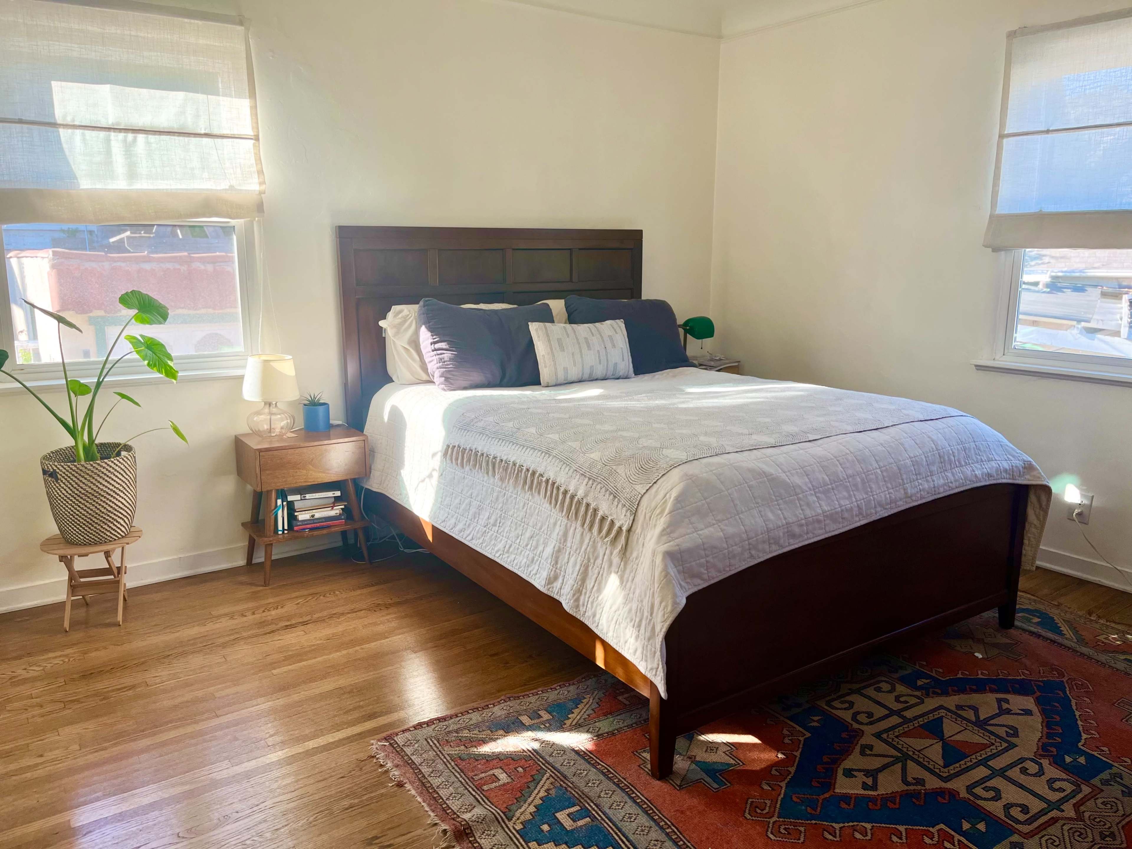 The image shows a tidy bedroom with a wooden bed, decorative pillows, a nightstand with a lamp, and a large area rug on the floor.