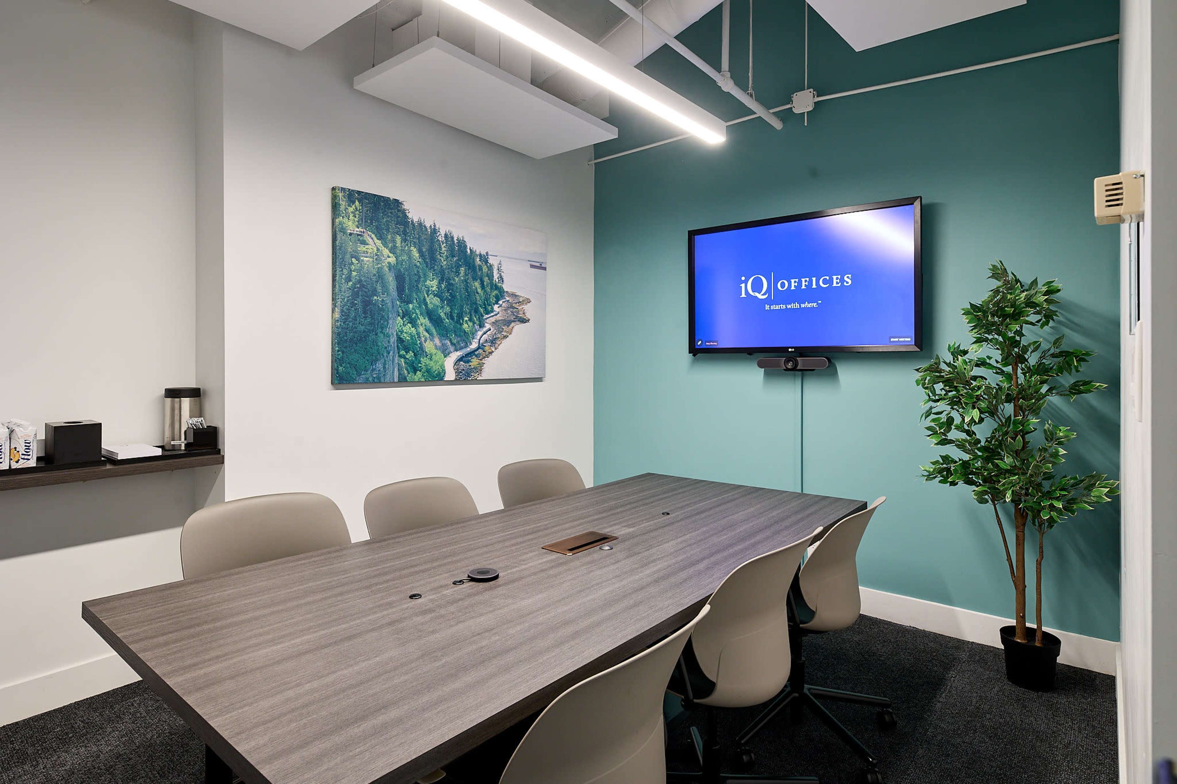 A conference room features a long table surrounded by chairs, a mounted television displaying a logo, and a green accent wall with a landscape painting.