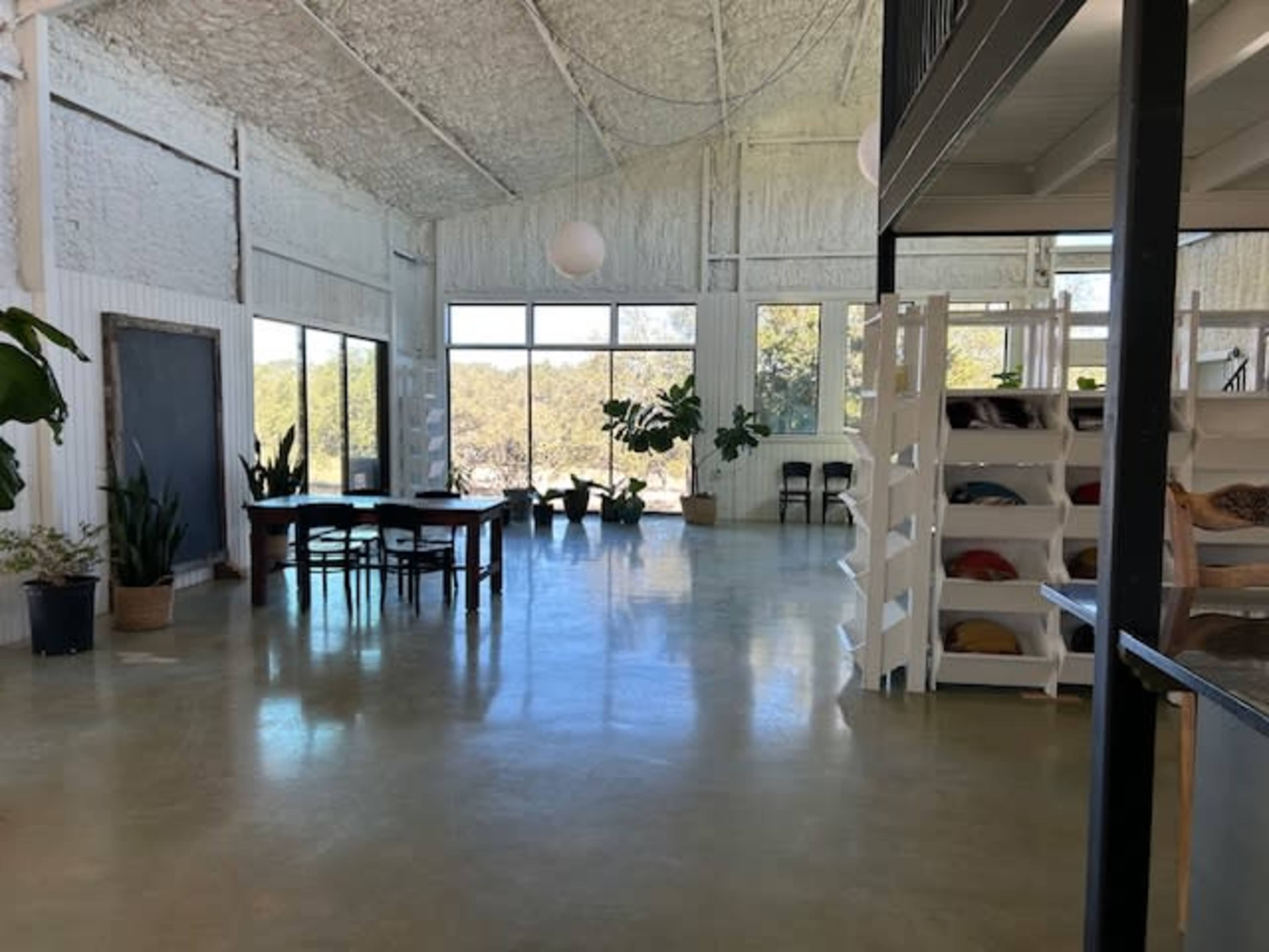 The image shows an open, airy space with large windows, a dining table, potted plants, and a shelving unit displaying cushions.