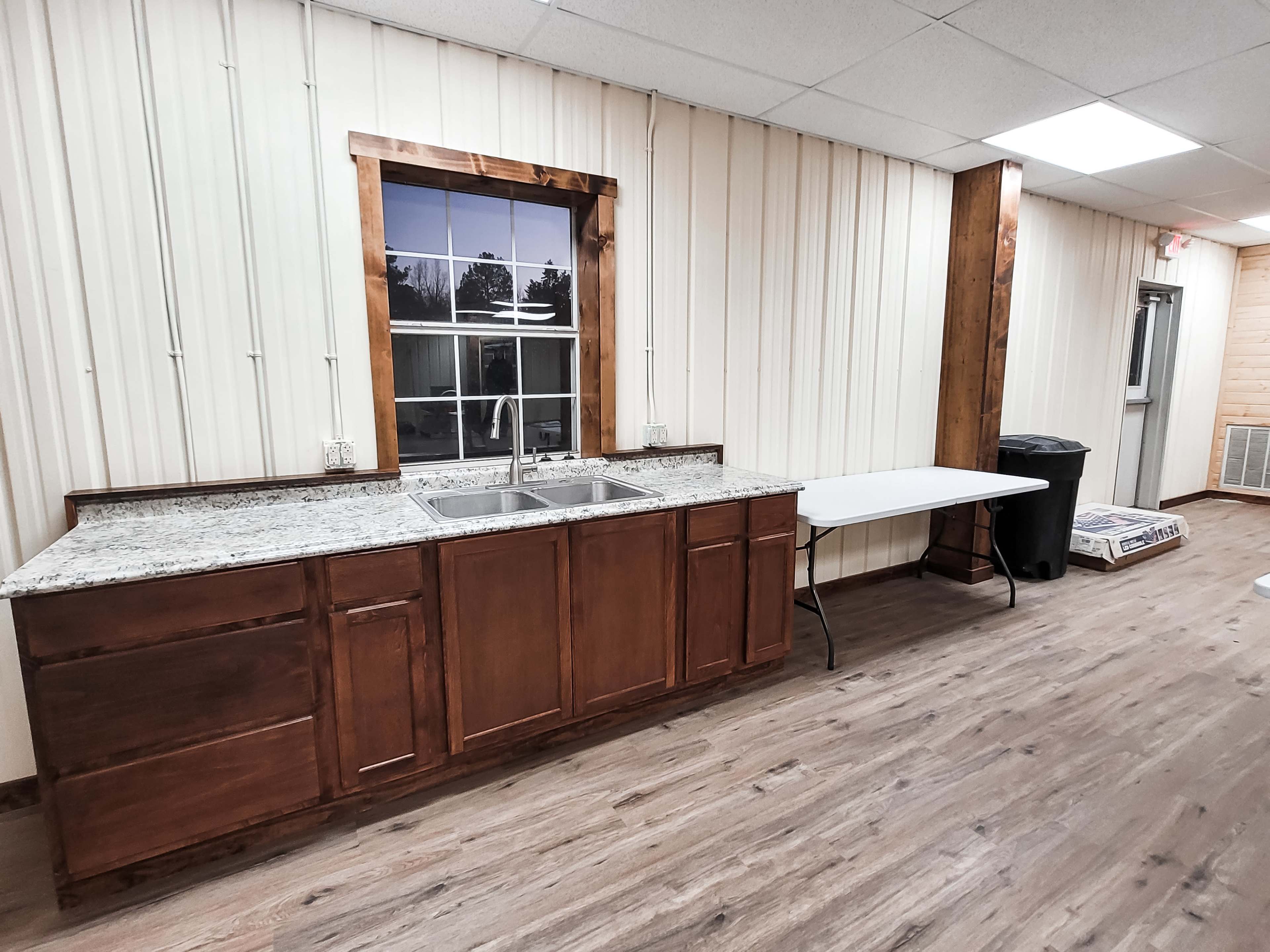 The image features a kitchen area with a wooden cabinet and granite countertop, a double sink beneath a window, and a white folding table next to a trash can.