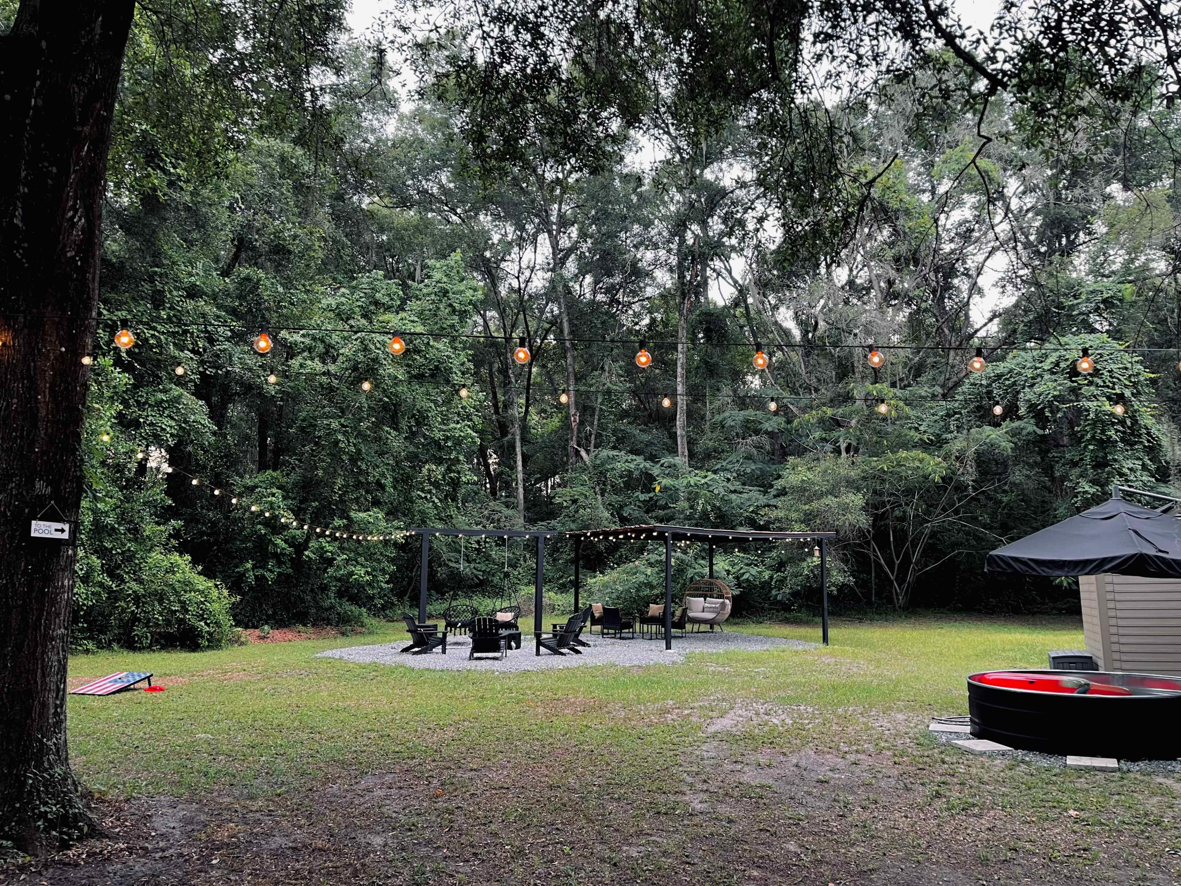 A grassy outdoor area surrounded by trees, featuring a seating arrangement with chairs and a canopy, illuminated by string lights.
