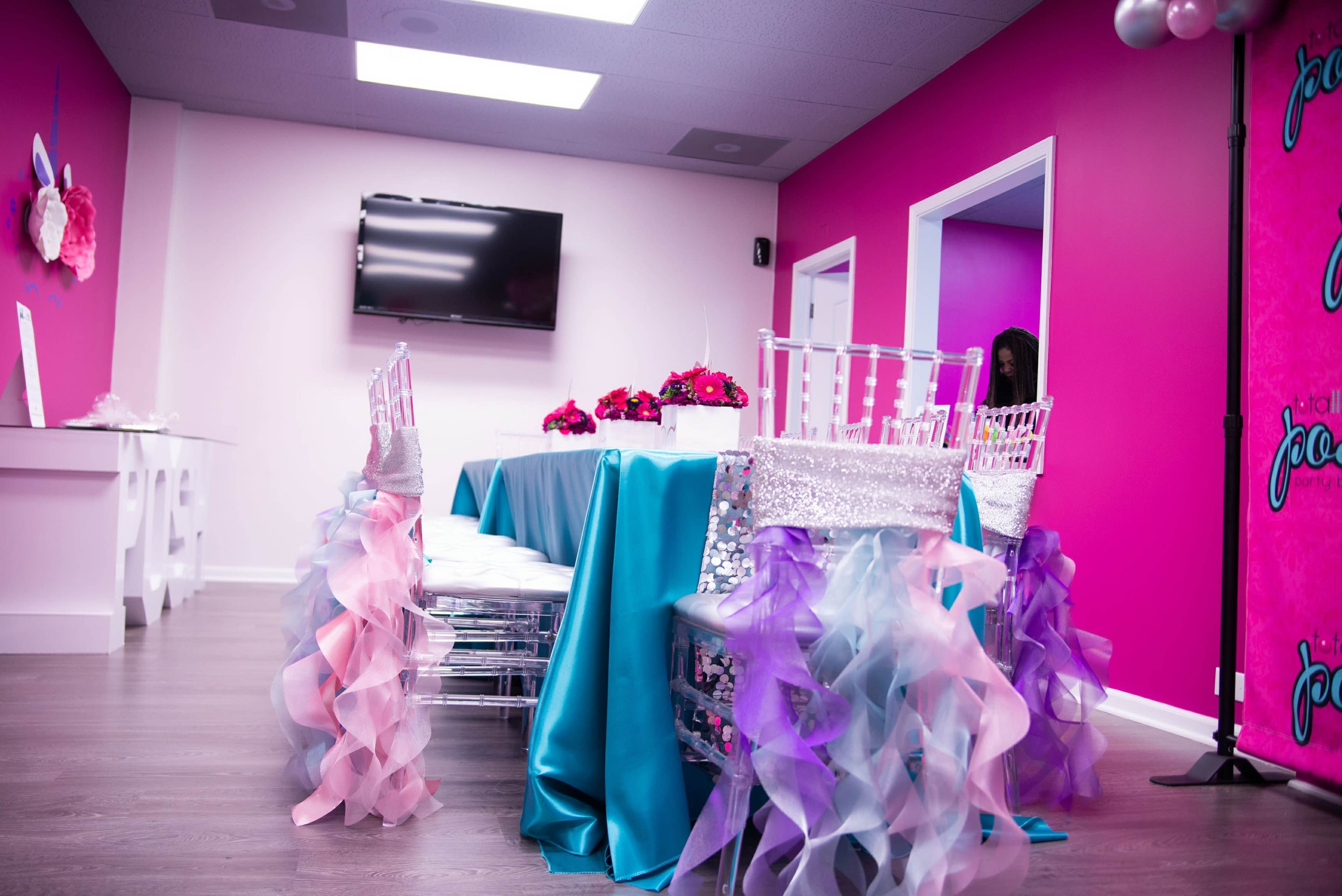 A brightly decorated room features a table set with a turquoise tablecloth and adorned with floral arrangements, while chairs draped in pastel ruffled fabric surround the table.