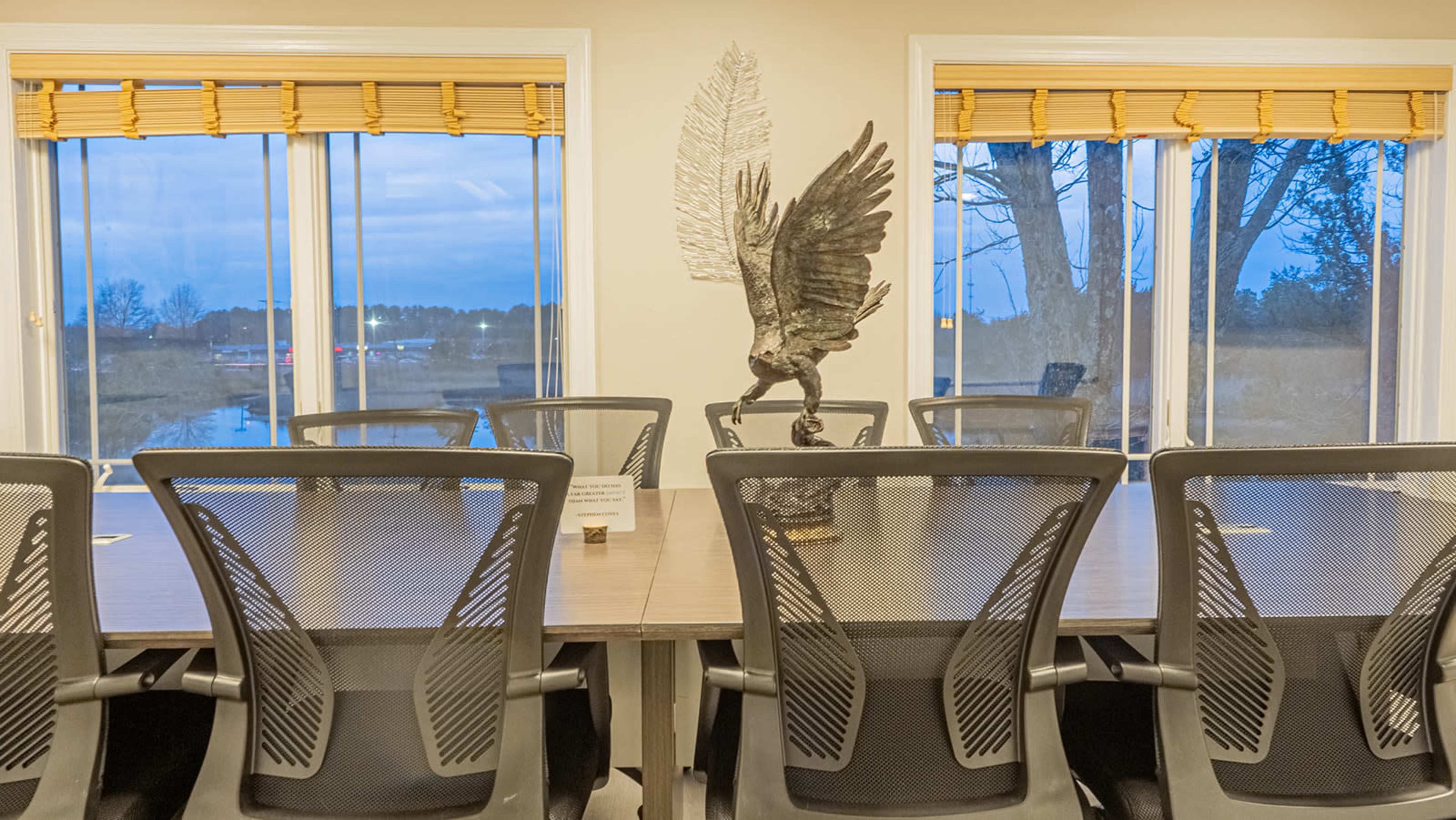 A conference room with a large table and mesh-backed chairs is illuminated by natural light coming through large windows, featuring a decorative eagle statue on the table.