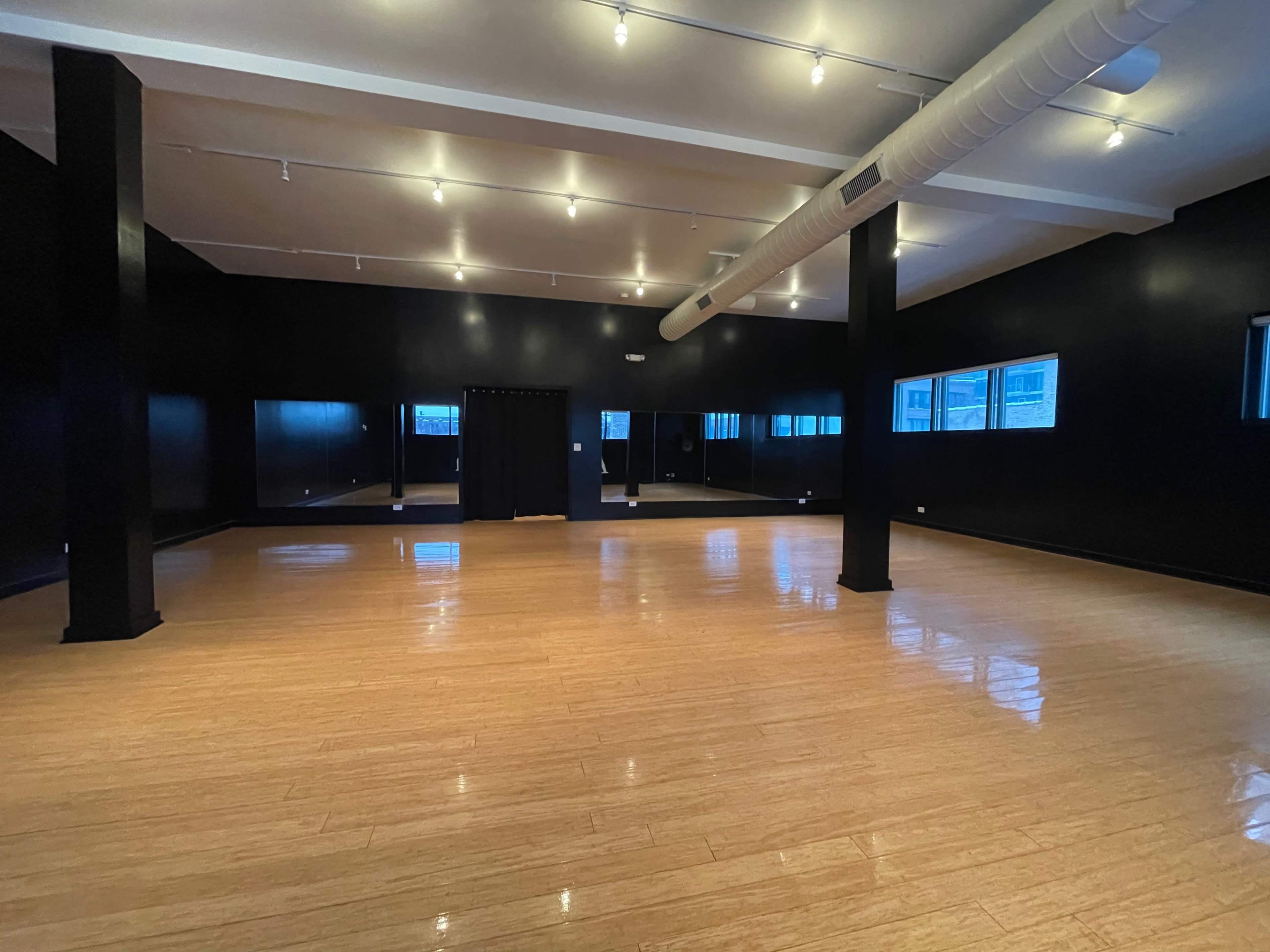 A spacious dance studio with a wooden floor, black walls, and large windows allowing natural light.