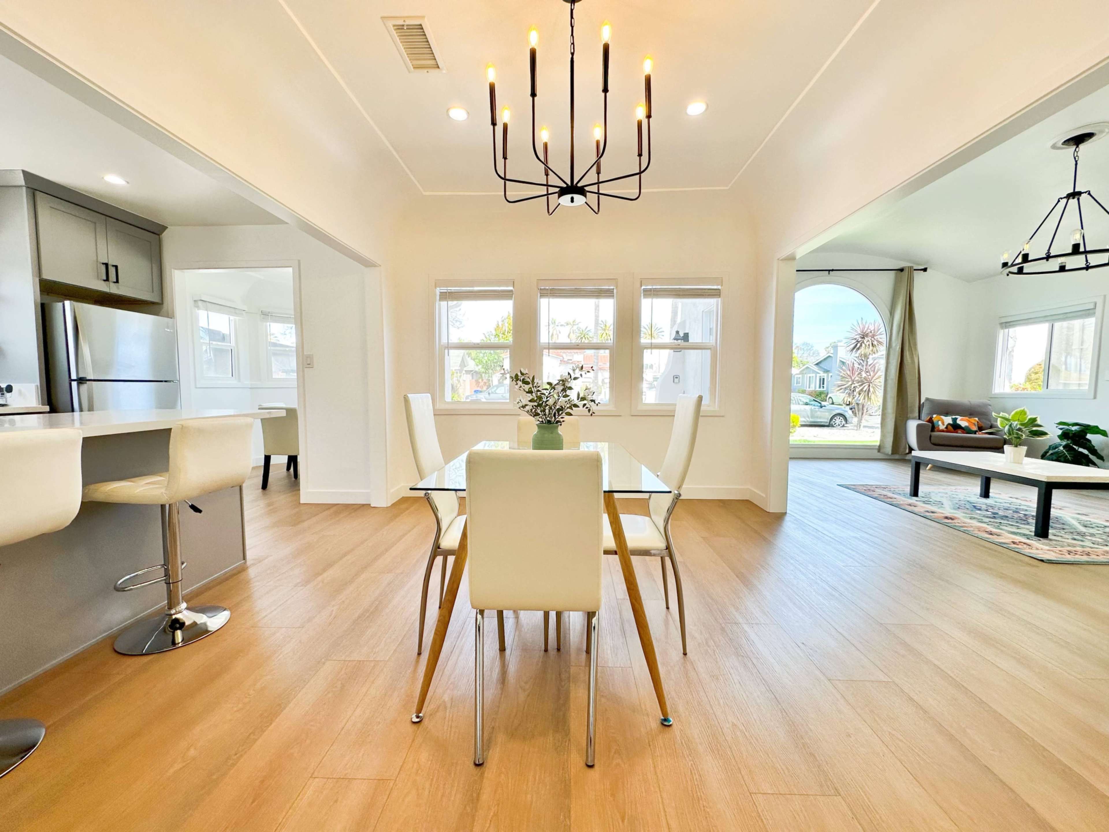 The image shows a modern dining area with a glass table surrounded by white chairs, complemented by a chandelier, and an open view to a living space with natural light.