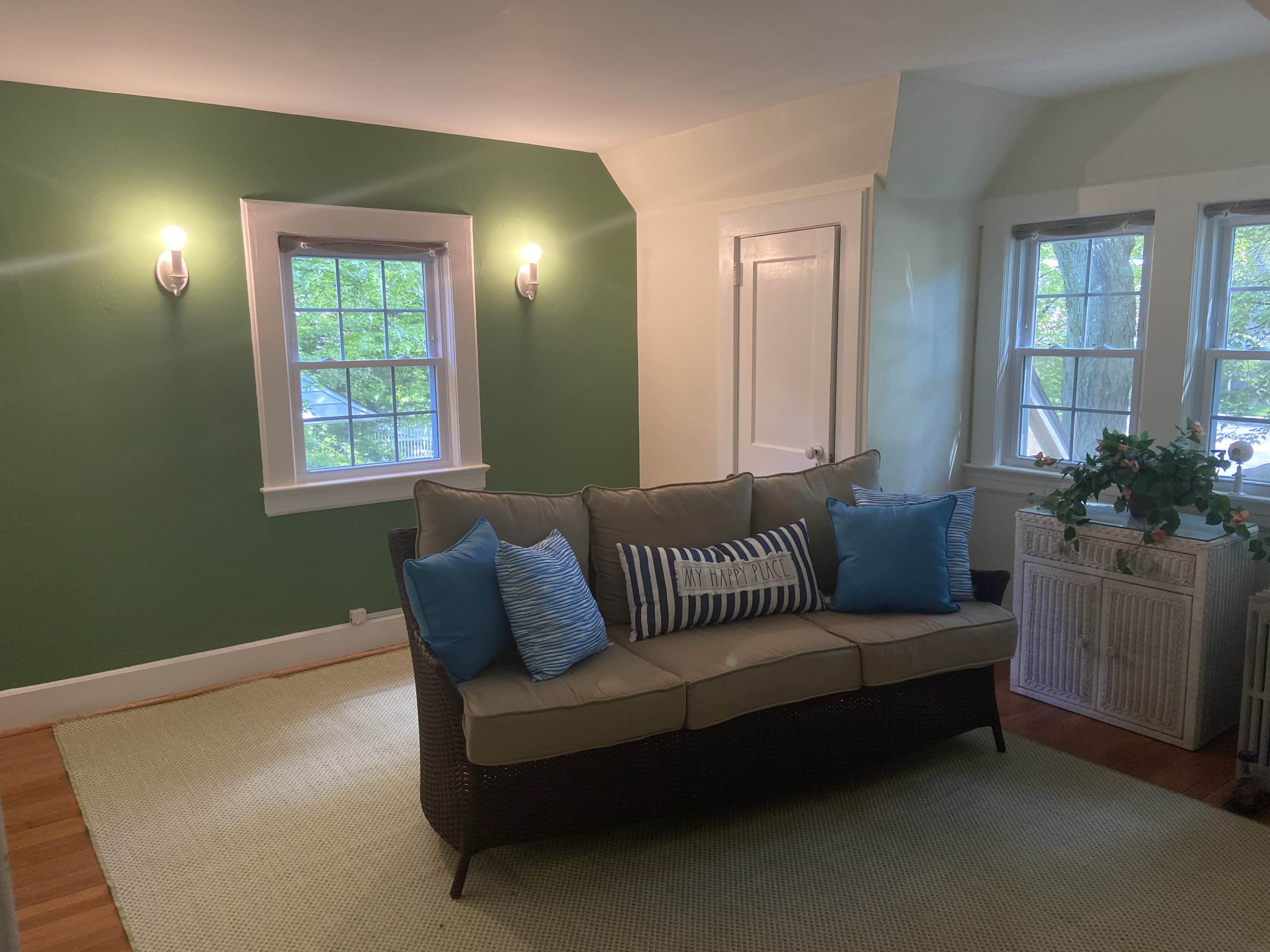 The scene shows a living room with a green accent wall, a light-colored sofa adorned with blue decorative pillows, and two windows allowing natural light to enter.