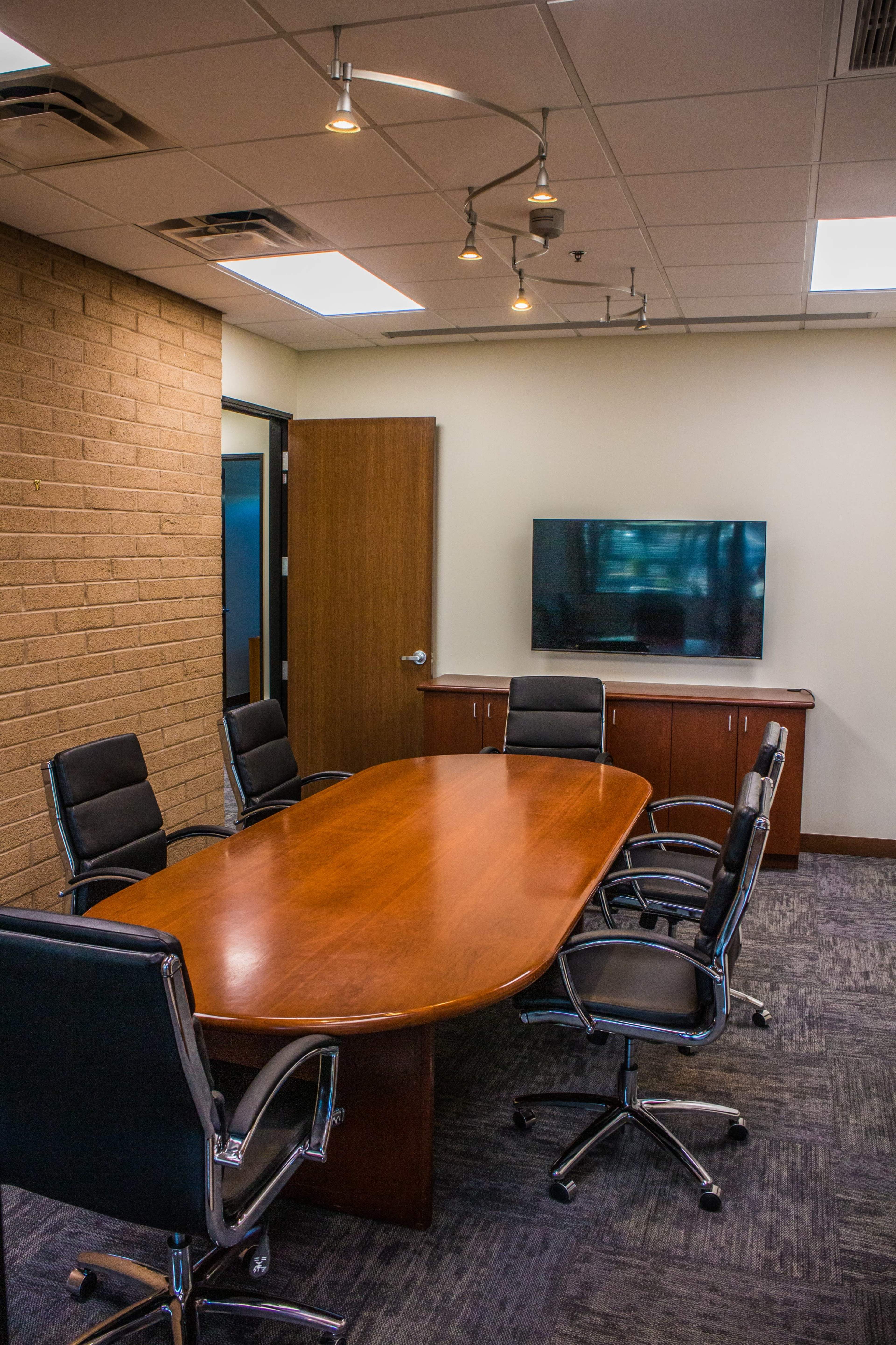 A conference room features a wooden oval table surrounded by black leather chairs, with a wall-mounted TV and a brick accent wall.