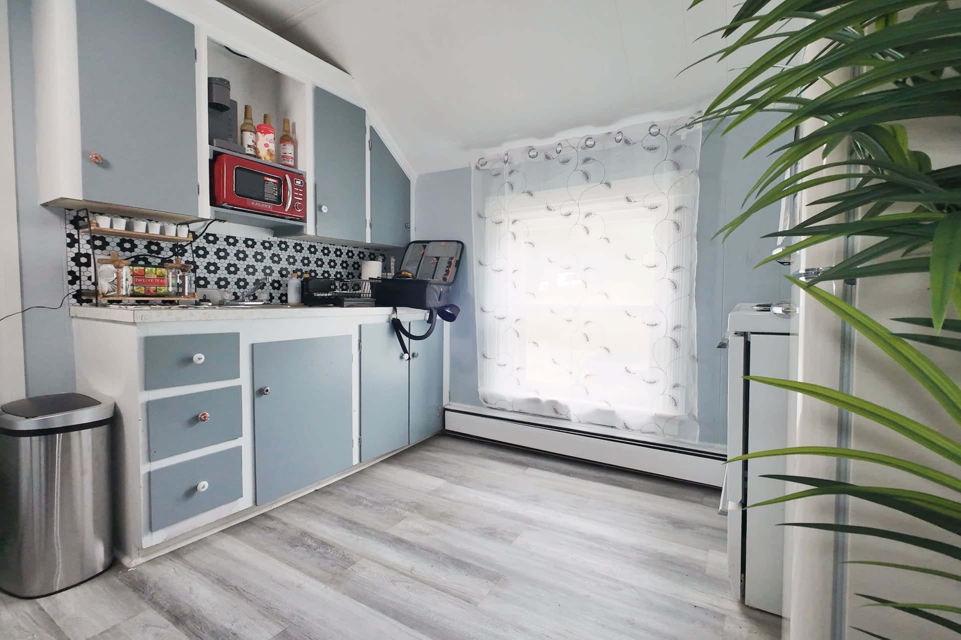 The image shows a bright kitchen with light blue walls, a white window adorned with sheer curtains, and cabinets displaying a variety of kitchen items.