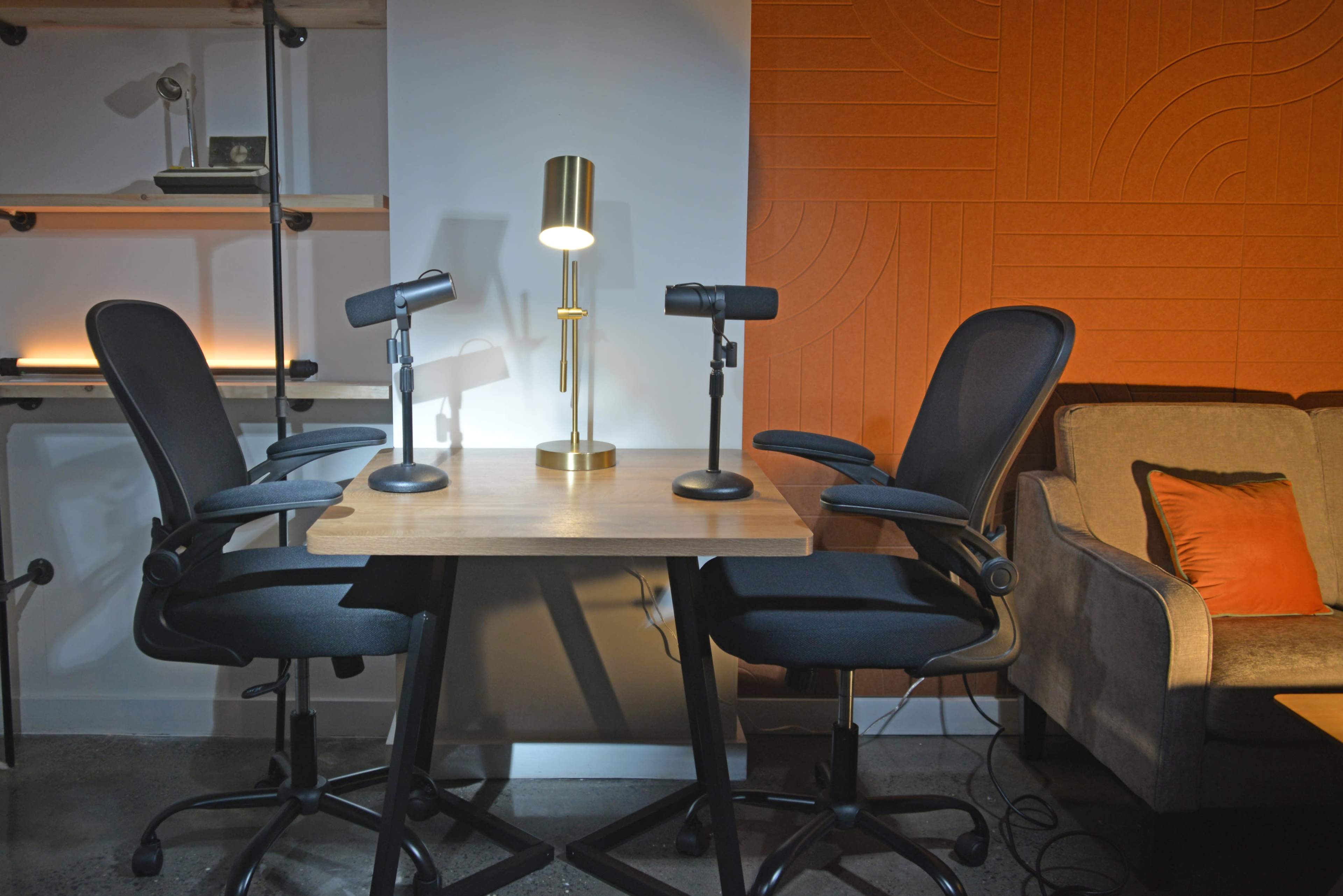The image shows two black office chairs positioned around a wooden table, with a gold lamp and two microphones on the table, against an orange wall with a textured design.