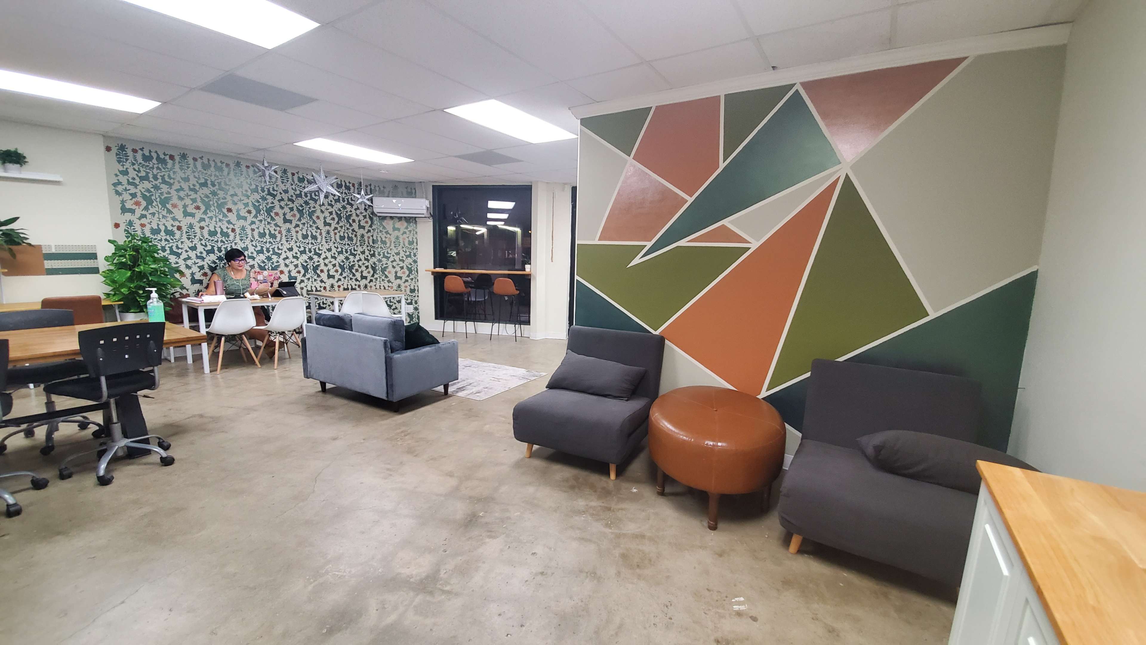 The image shows a modern, open office space with a colorful geometric wall, seating areas, and a dining table where one person is working.