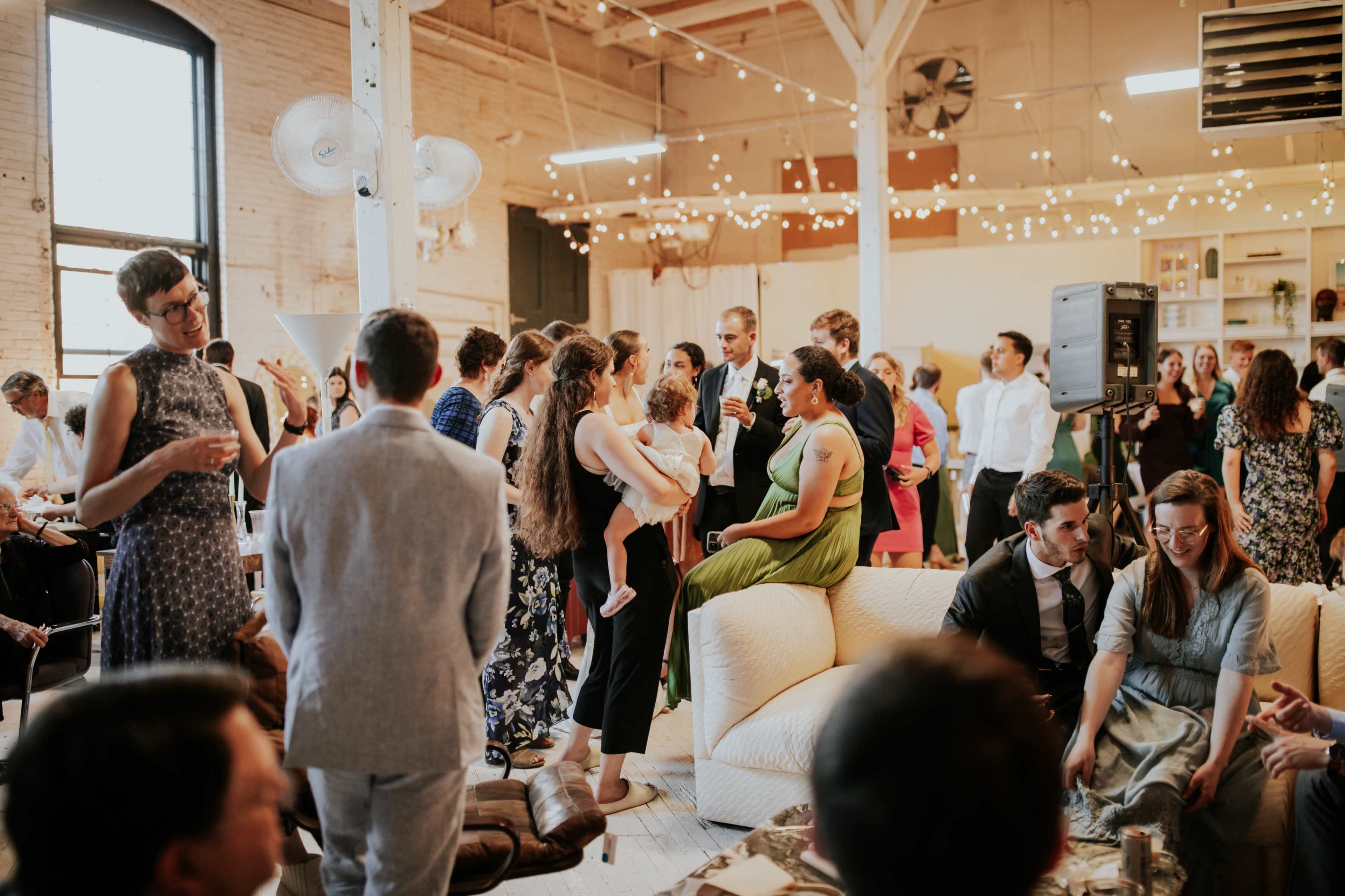 A large group of people socialize at a lively indoor event, surrounded by string lights and various seating arrangements.