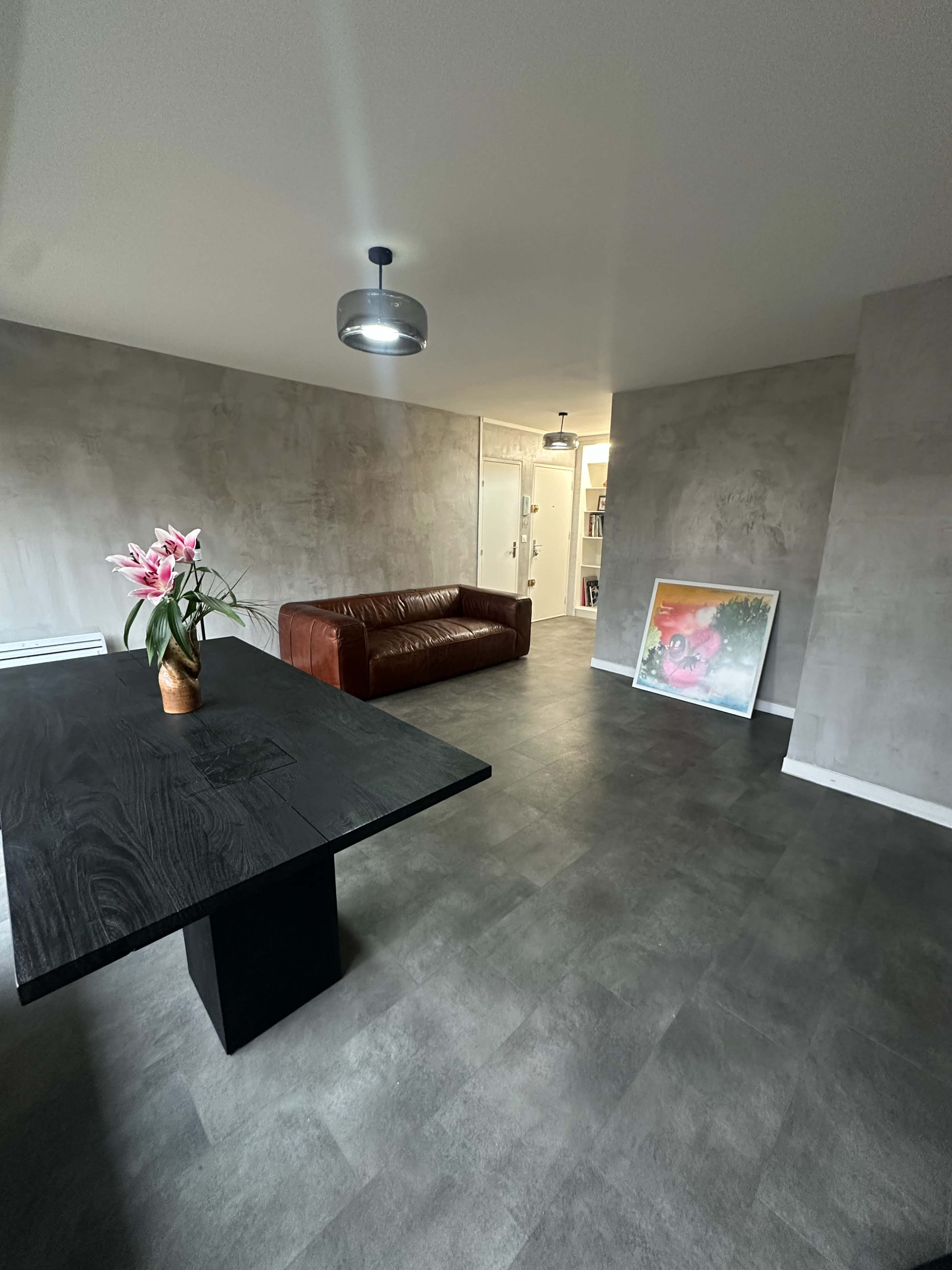 The image shows a modern interior space with a brown leather sofa, a large black dining table, and a colorful painting leaning against the wall.