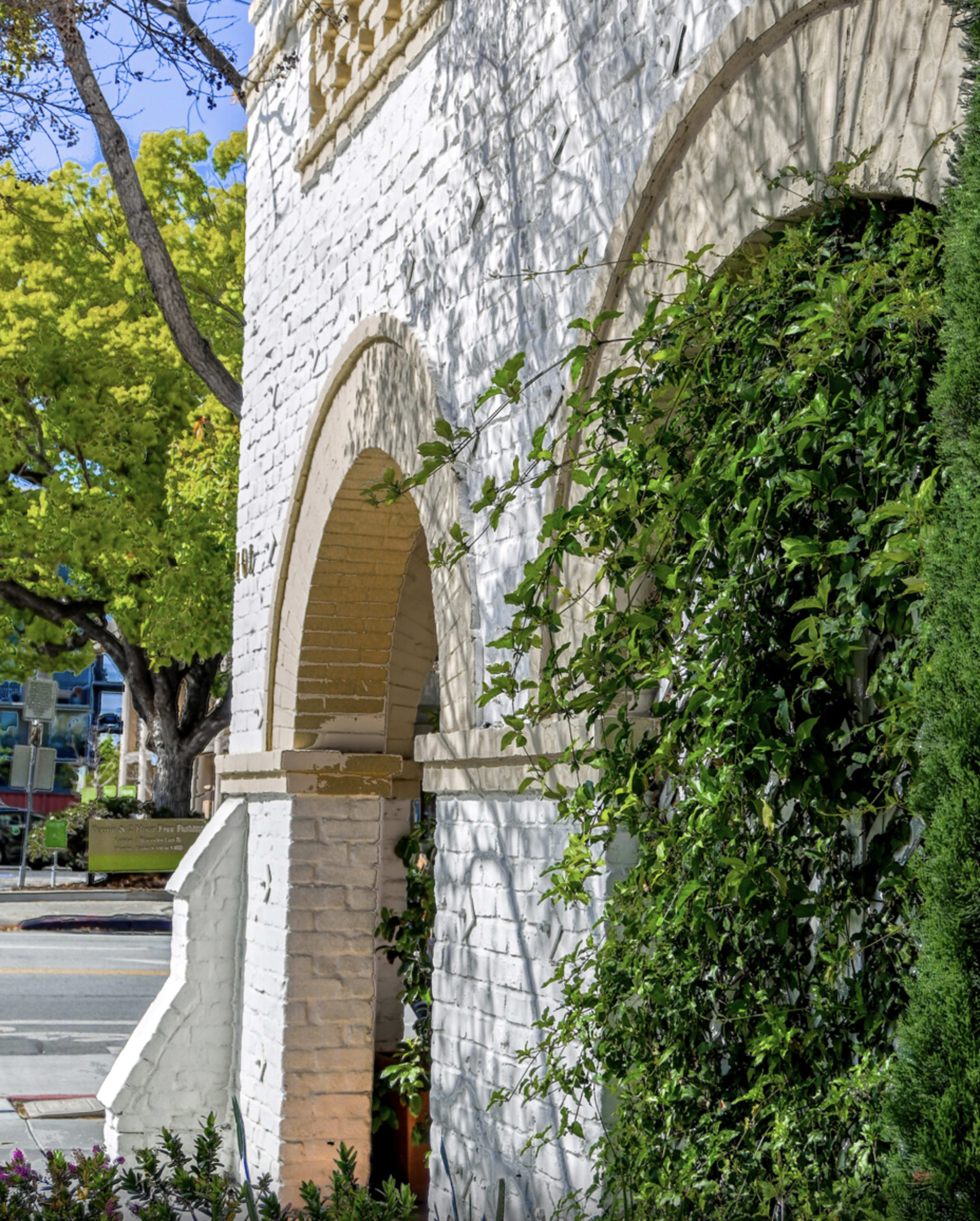 A white brick building with arched openings, surrounded by green foliage and trees.