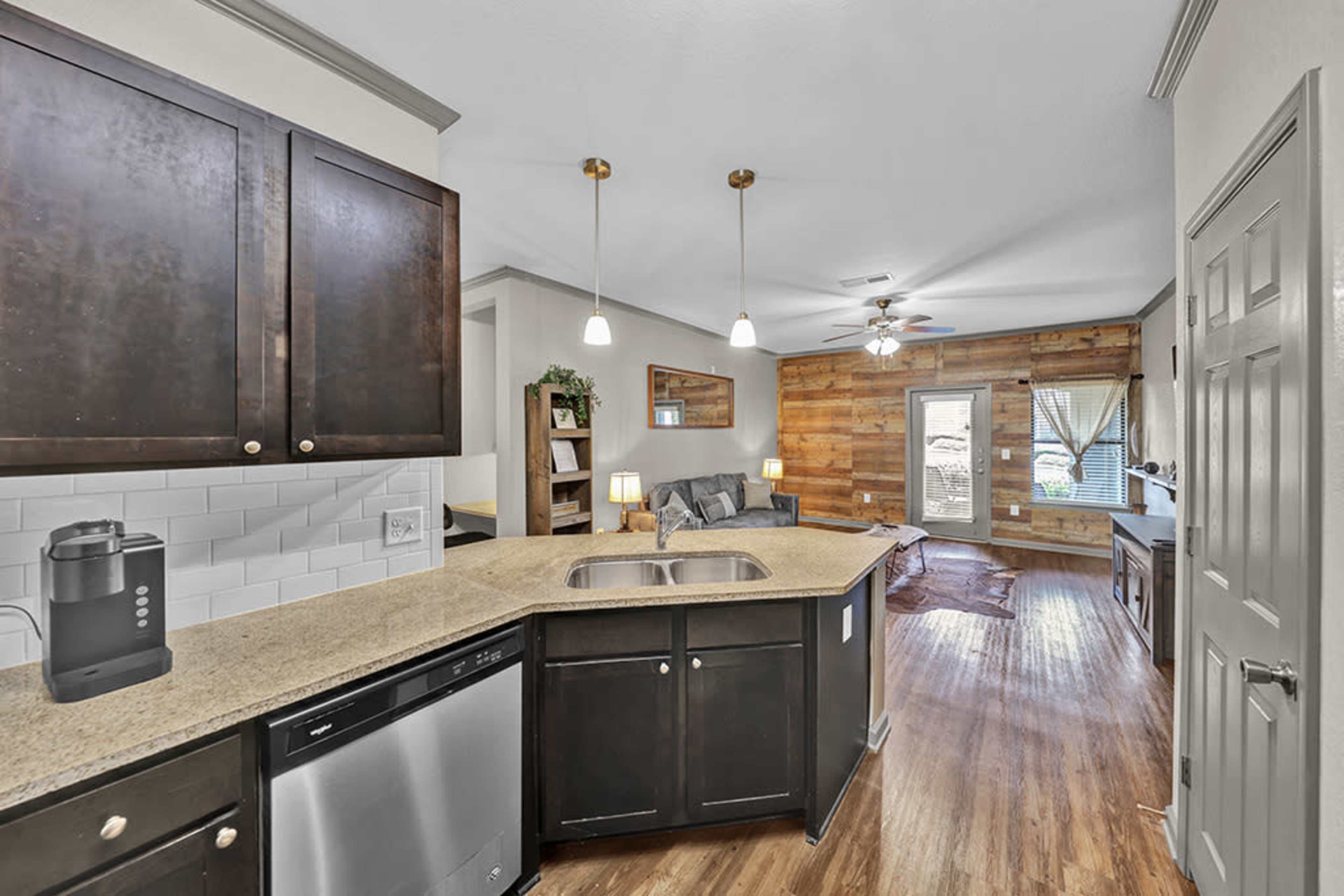 A kitchen with dark cabinets and a granite countertop, adjacent to a living area featuring a wooden accent wall and a door leading outside.