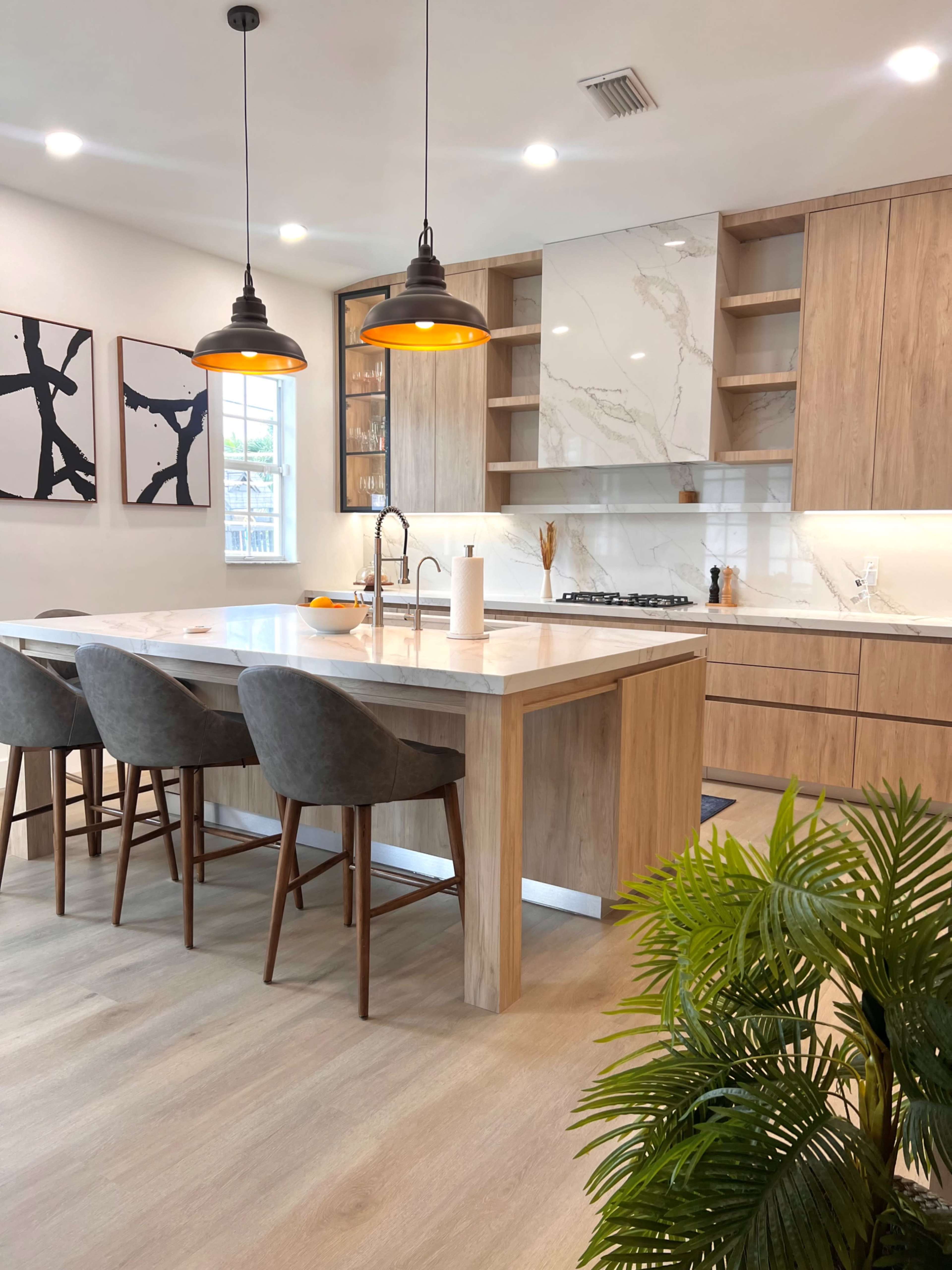 A modern kitchen features a large wooden island with four gray upholstered chairs, pendant lights overhead, and minimalist cabinetry alongside a marble backsplash.