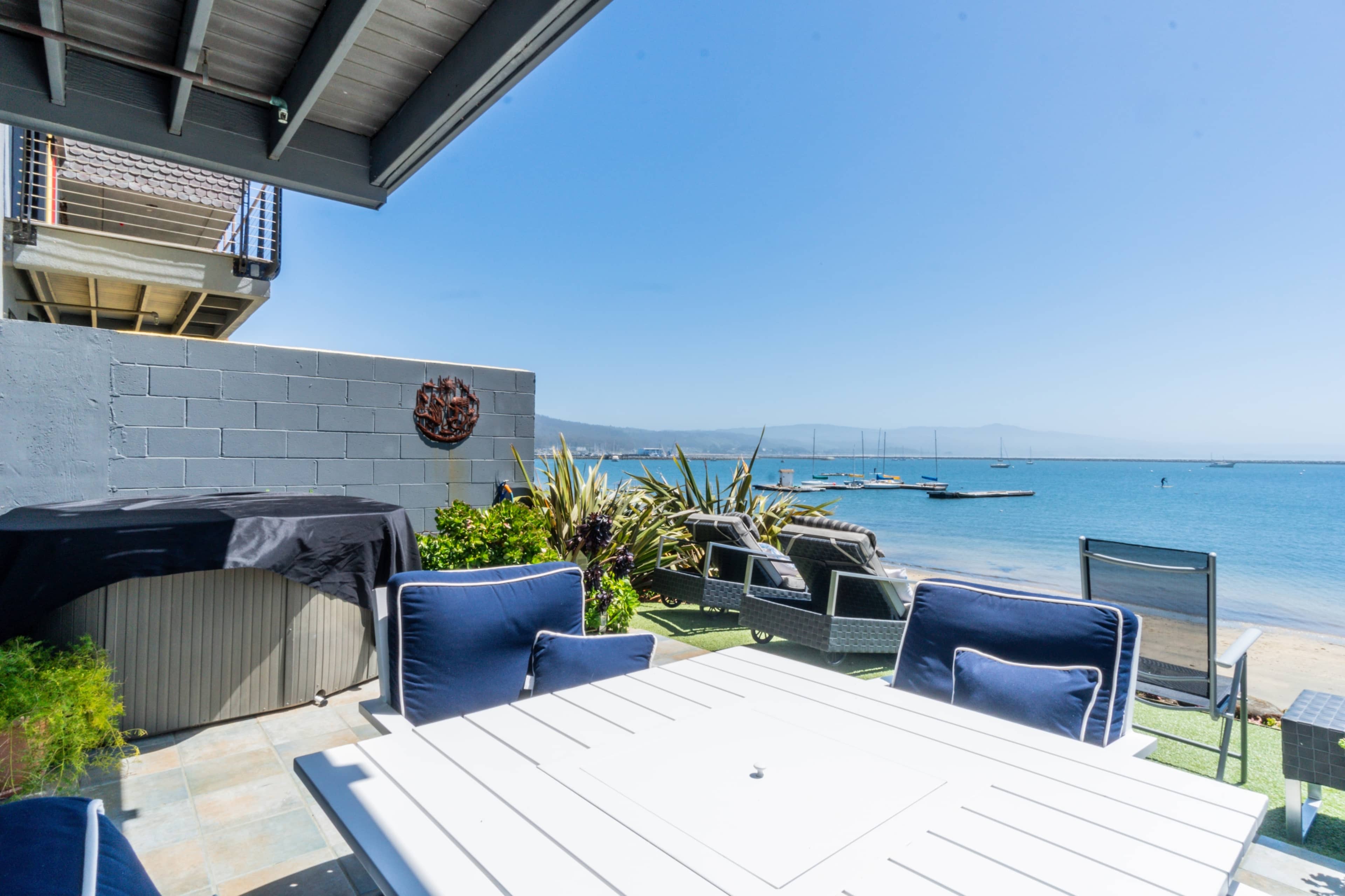 The image shows a patio area with a white table and chairs overlooking a calm bay with boats in the distance.