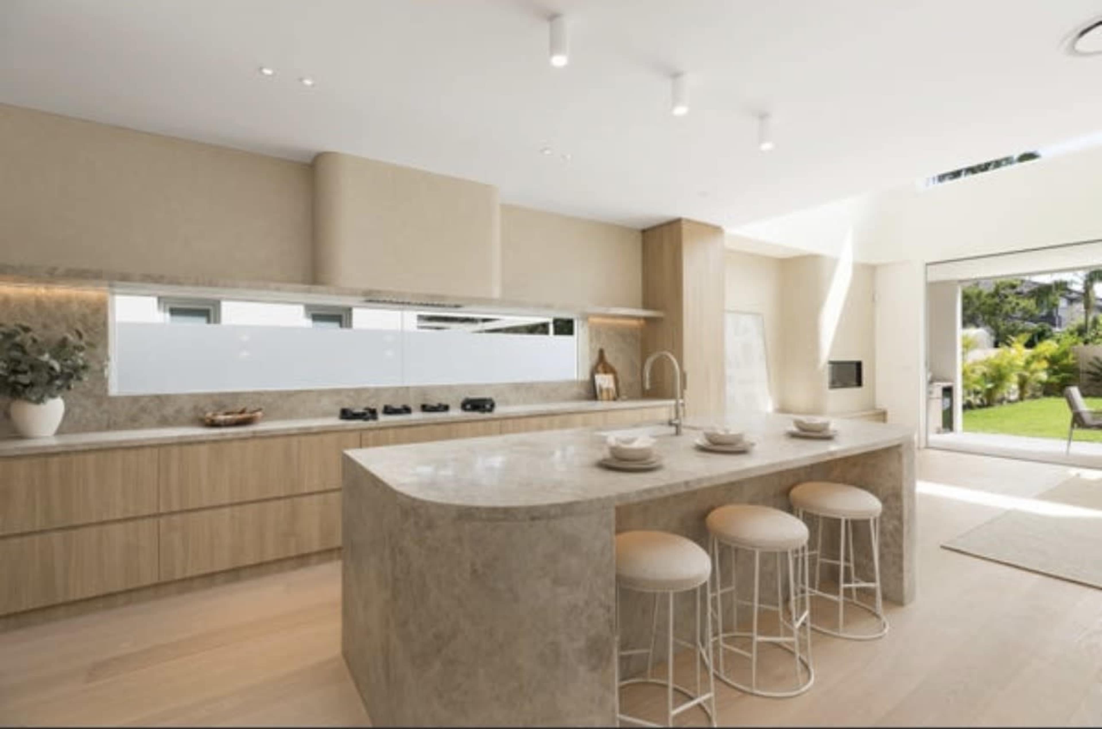 The kitchen features a modern design with light wood cabinets, a large stone island, and bar stools, alongside an open view to an outdoor area.