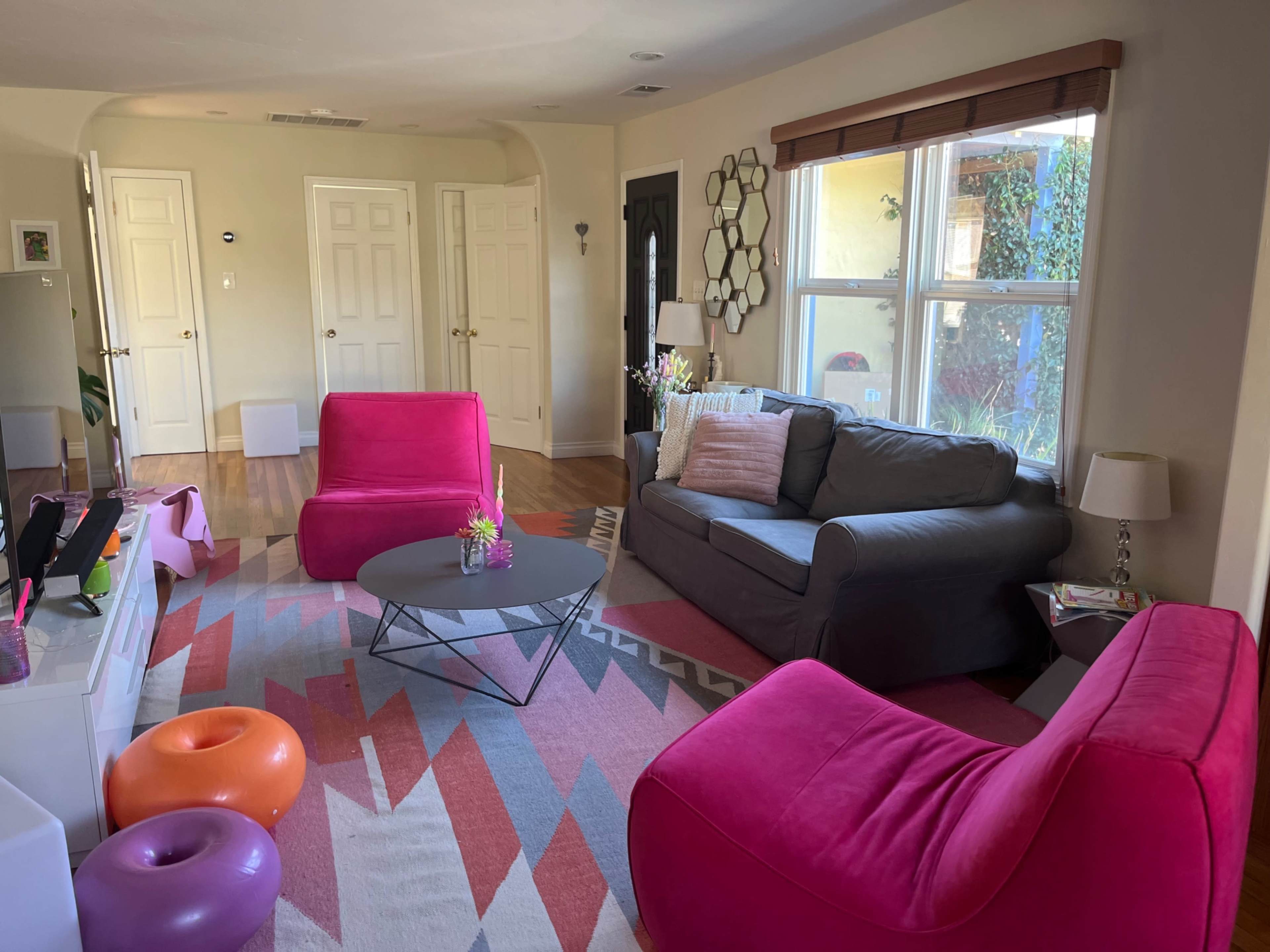 The living room features a gray sofa and two bright pink lounge chairs, arranged around a round coffee table on a patterned area rug.
