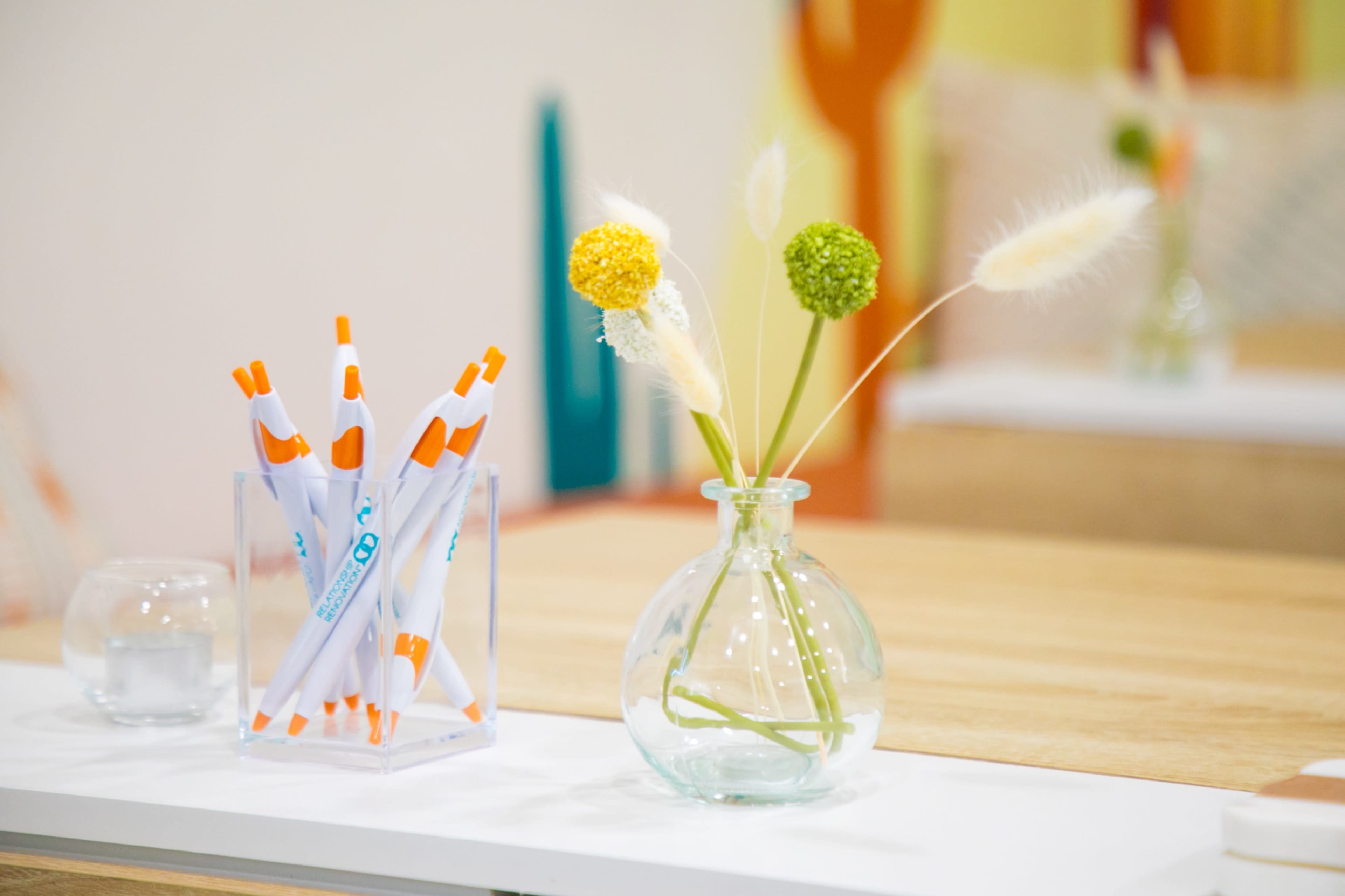 A clear vase with decorative flowers sits next to a container of white and orange markers on a wooden tabletop.