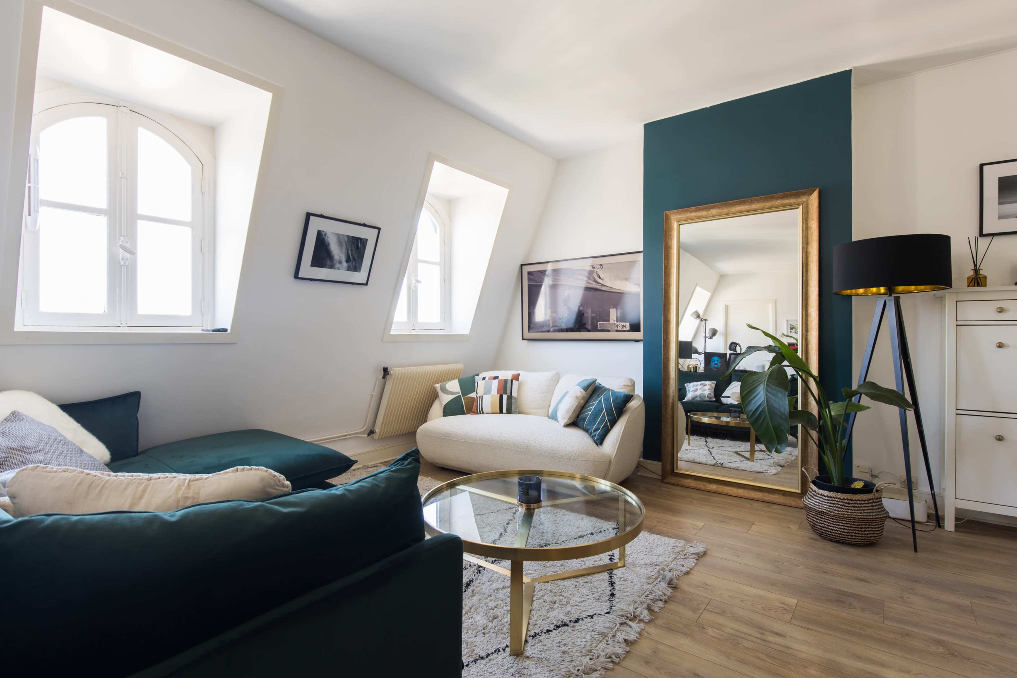 A modern living room features a curved sofa, a coffee table, and a large mirror against a colorful accent wall, with natural light streaming through angled windows.