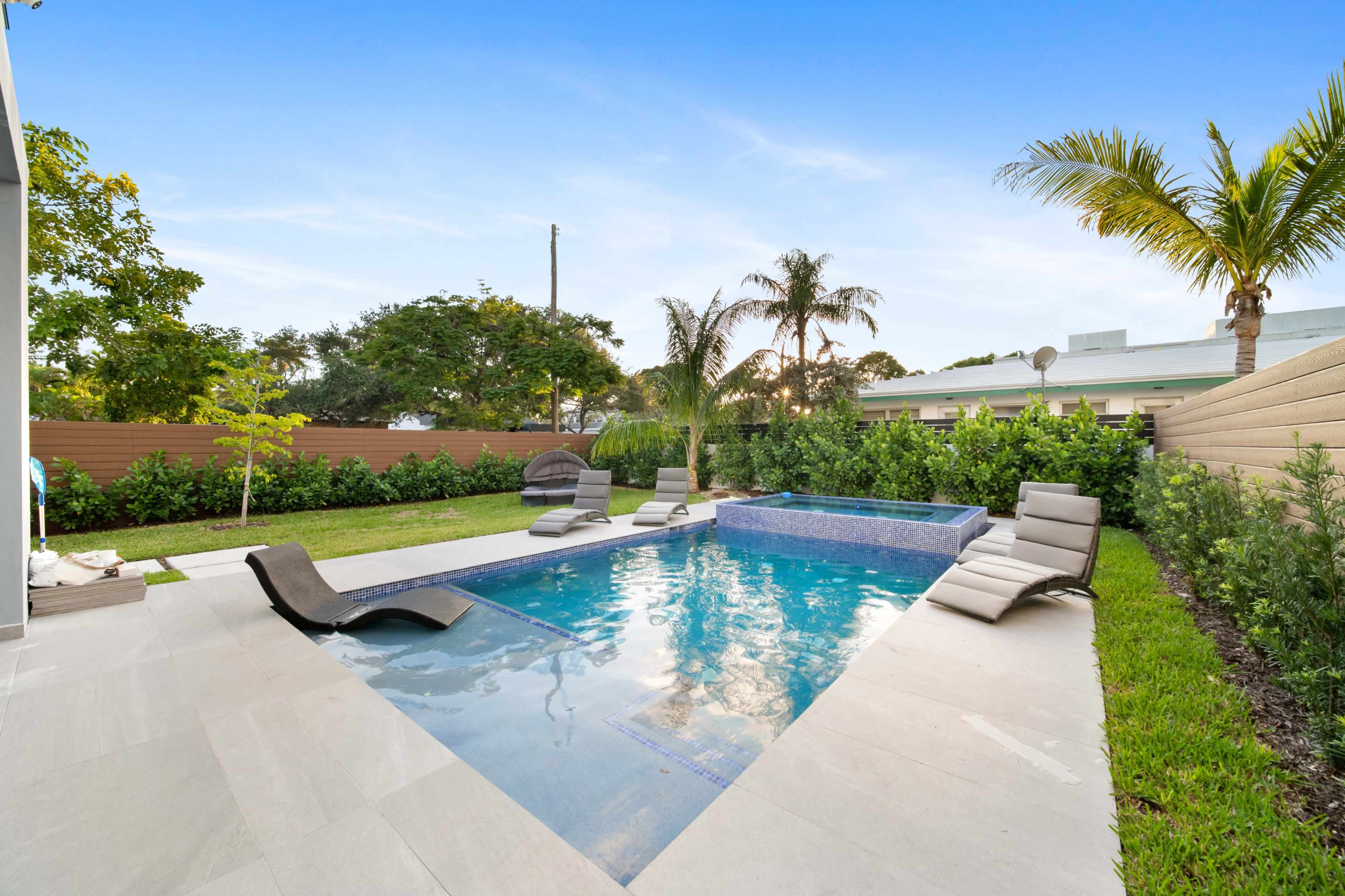A backyard features a swimming pool with a spa and several lounge chairs surrounded by palm trees and manicured grass.