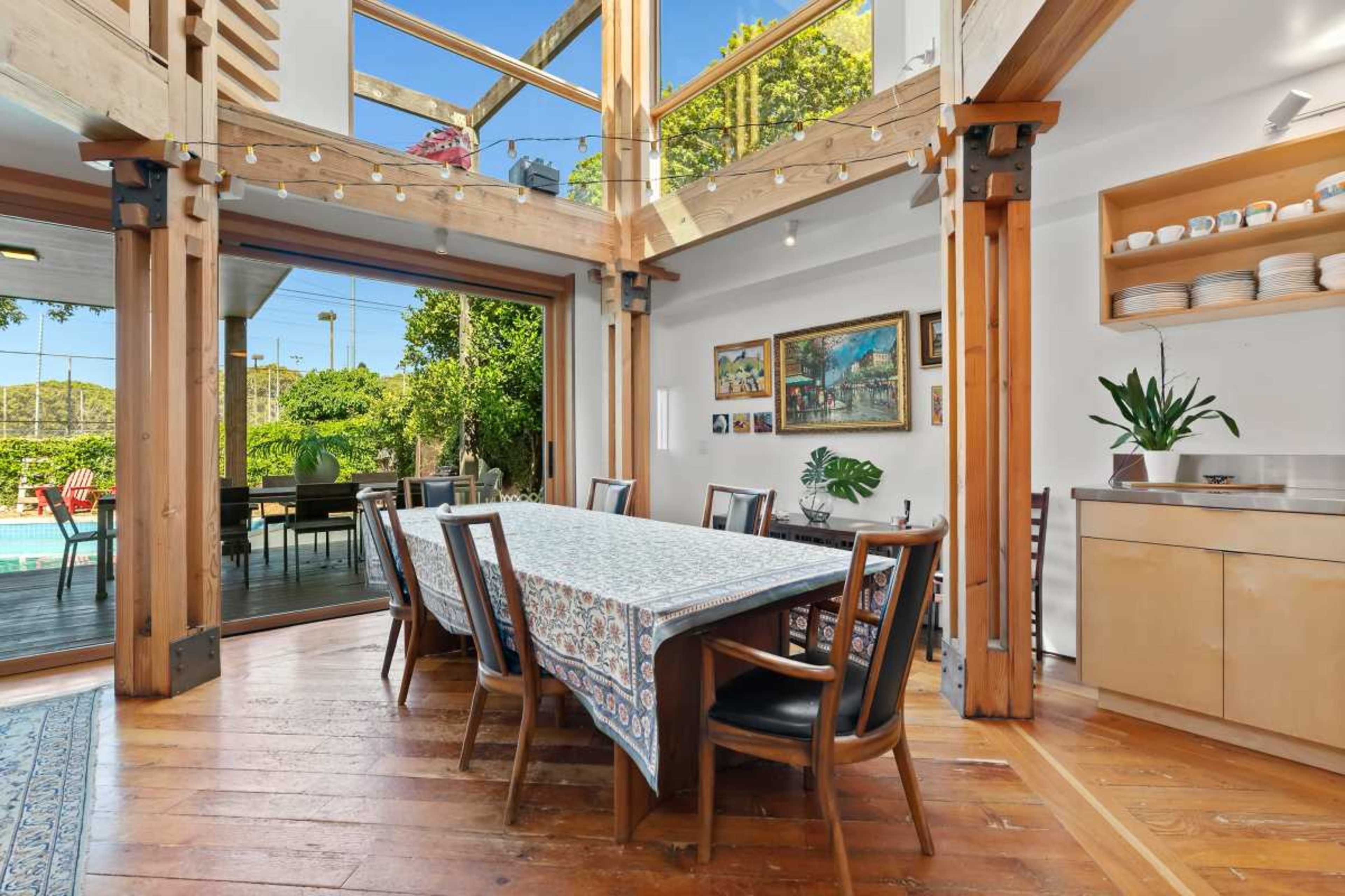 The image depicts a dining area with a large wooden table, surrounded by chairs, and large windows that open to an exterior view of greenery and a pool.
