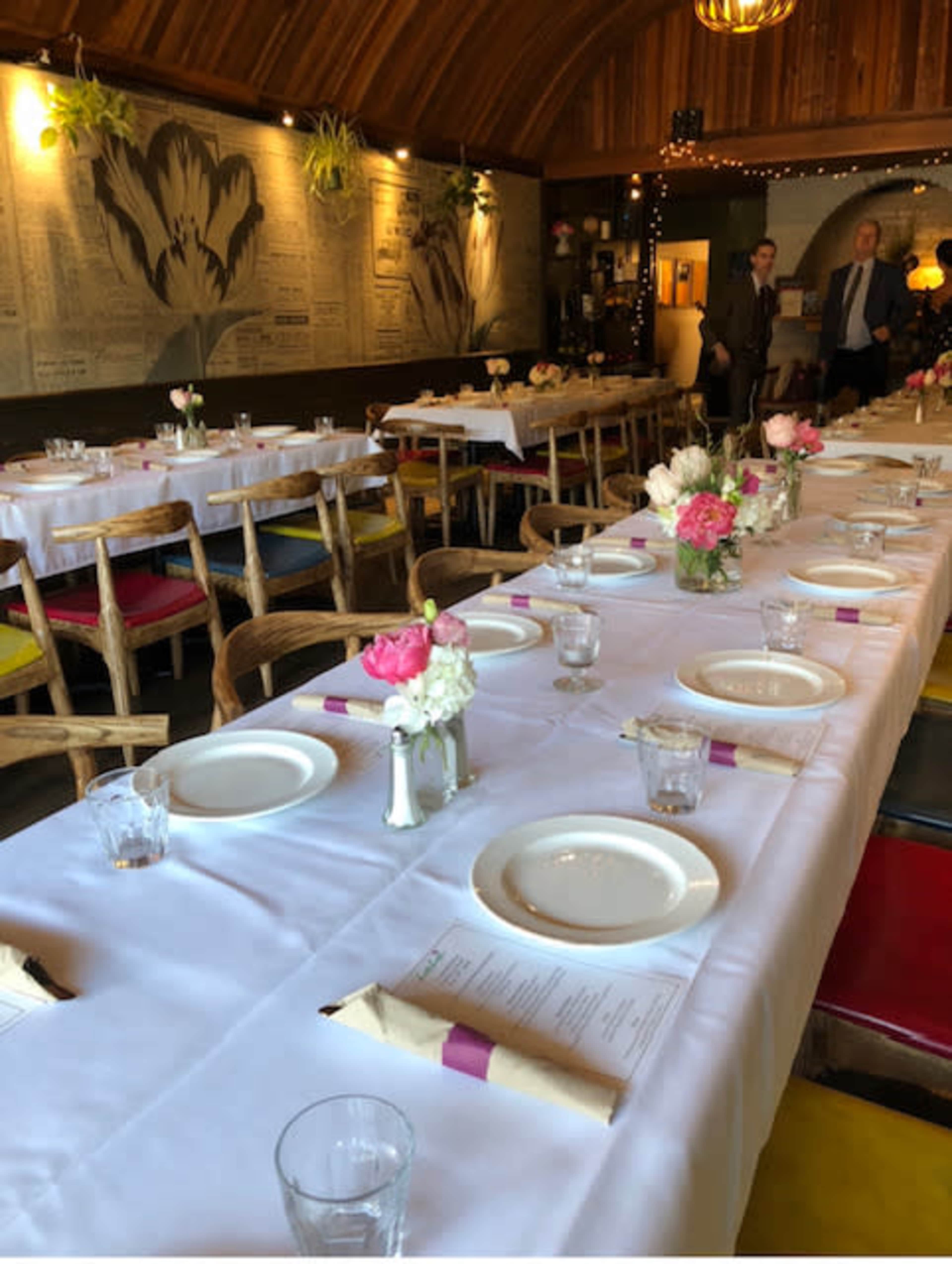 A long, elegantly set dining table with white tablecloths, flower centerpieces, and place settings is arranged in a quaint restaurant with a wood-paneled interior.