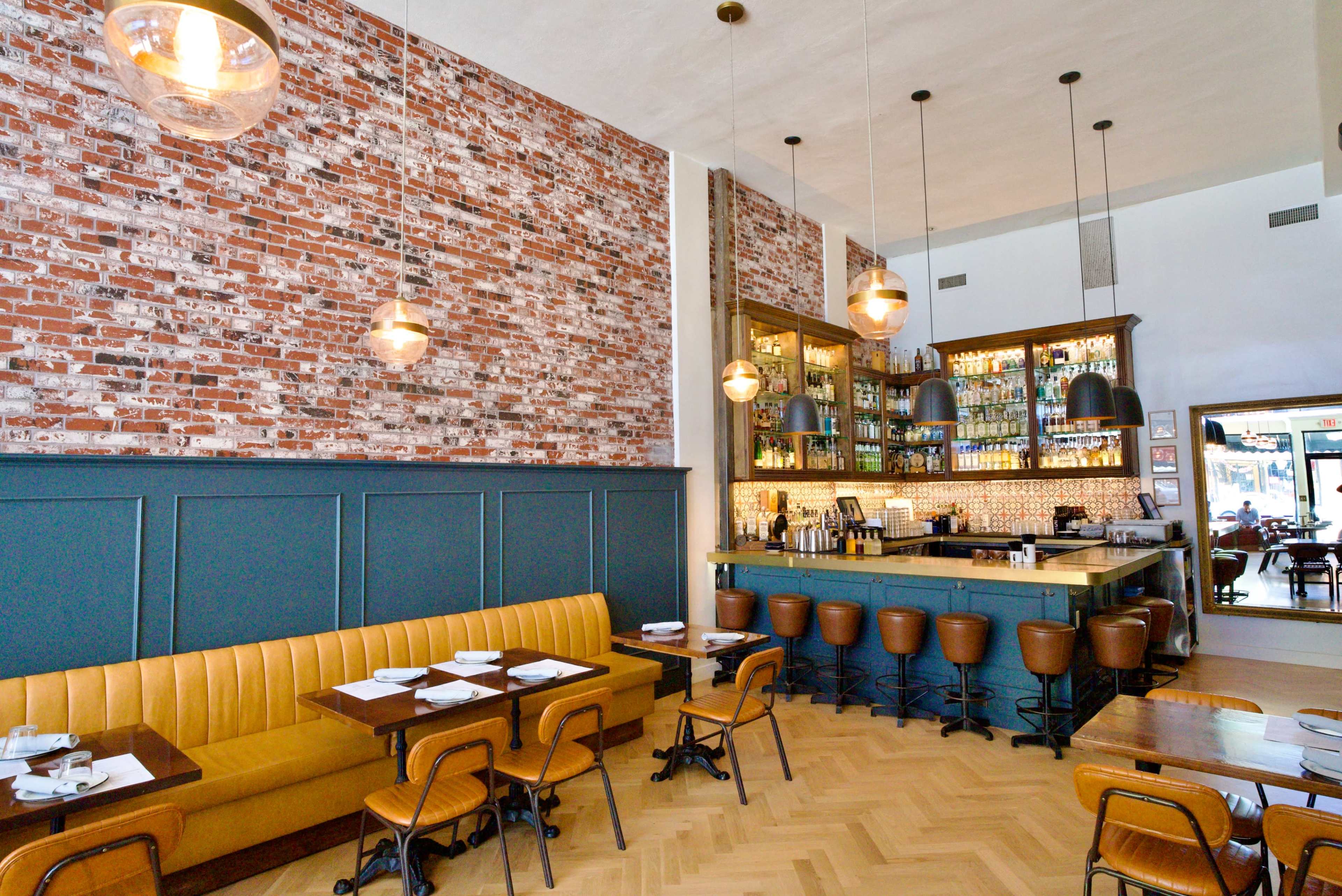 The image shows a modern restaurant interior with exposed brick walls, wooden tables, and a bar area featuring shelves of bottles.