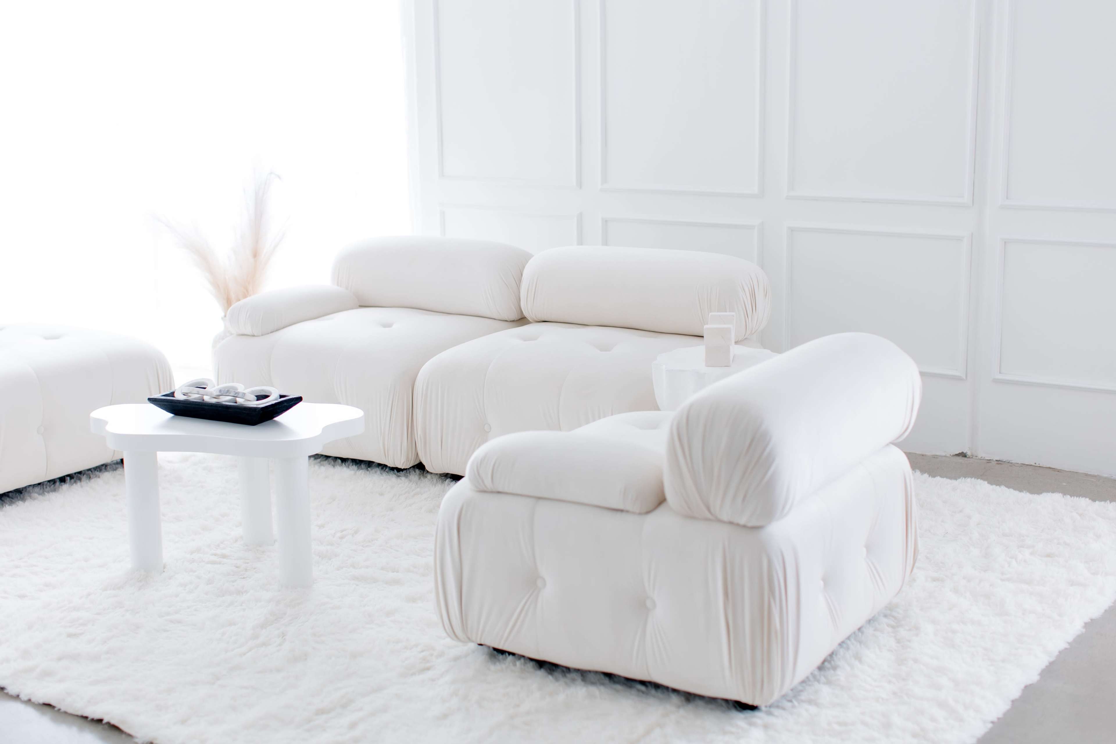 A modern, white sectional sofa is positioned on a plush, white area rug in a bright, minimalistic room.