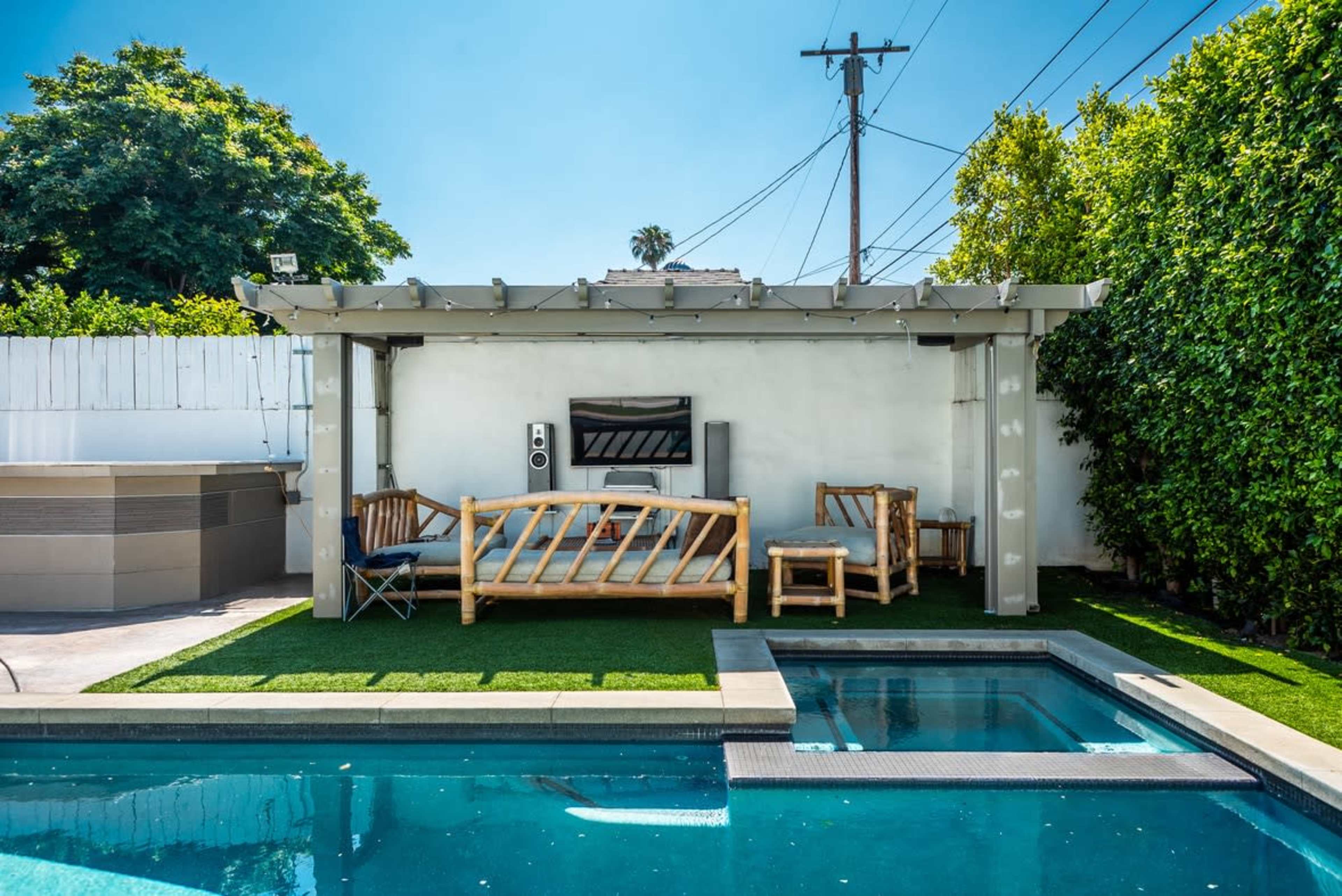 A modern outdoor seating area with a TV is situated beside a swimming pool, surrounded by greenery and powered by string lights.