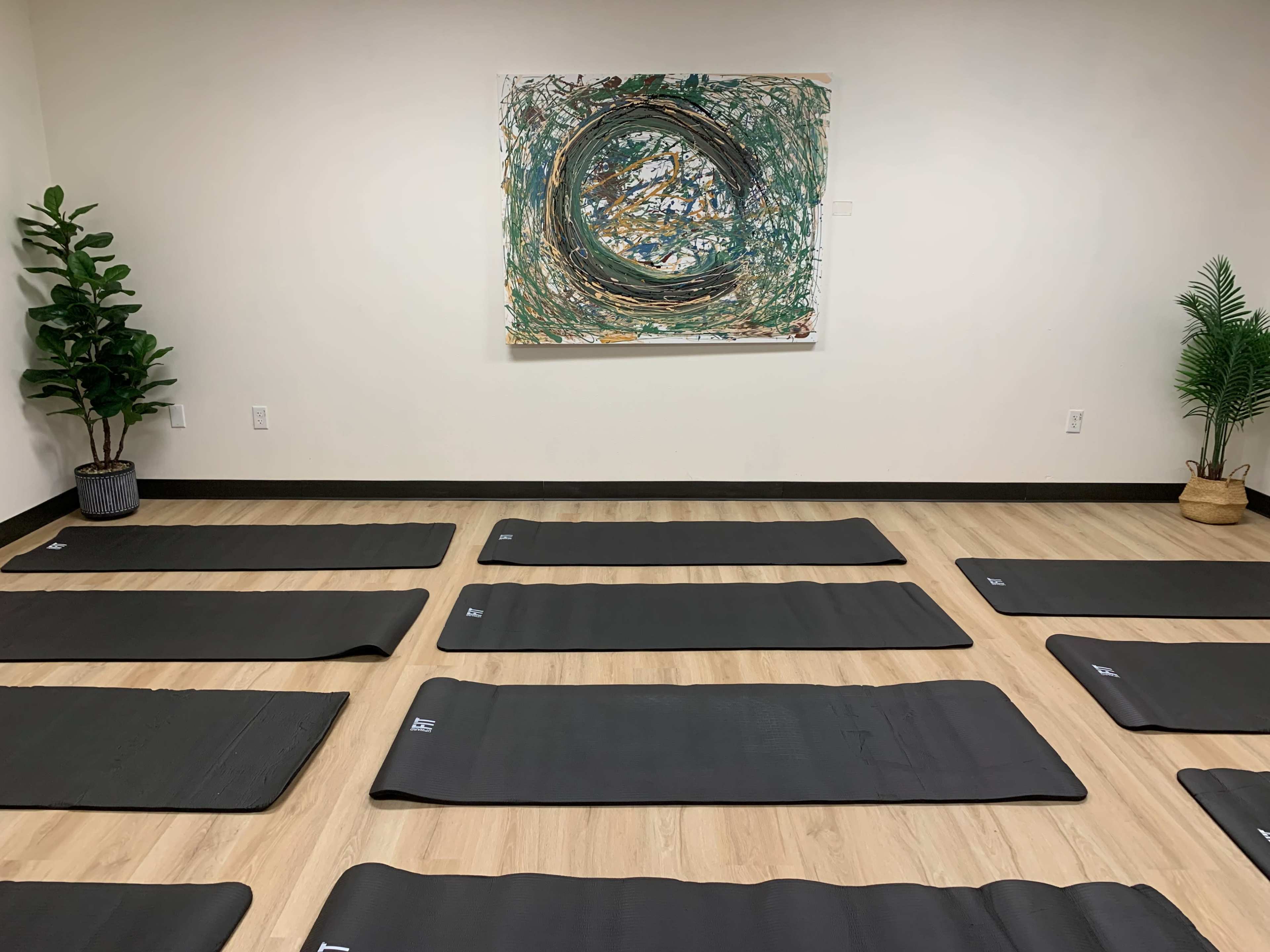 The image shows a yoga studio with numerous black mats arranged on the wooden floor, and a circular abstract painting hanging on the wall.