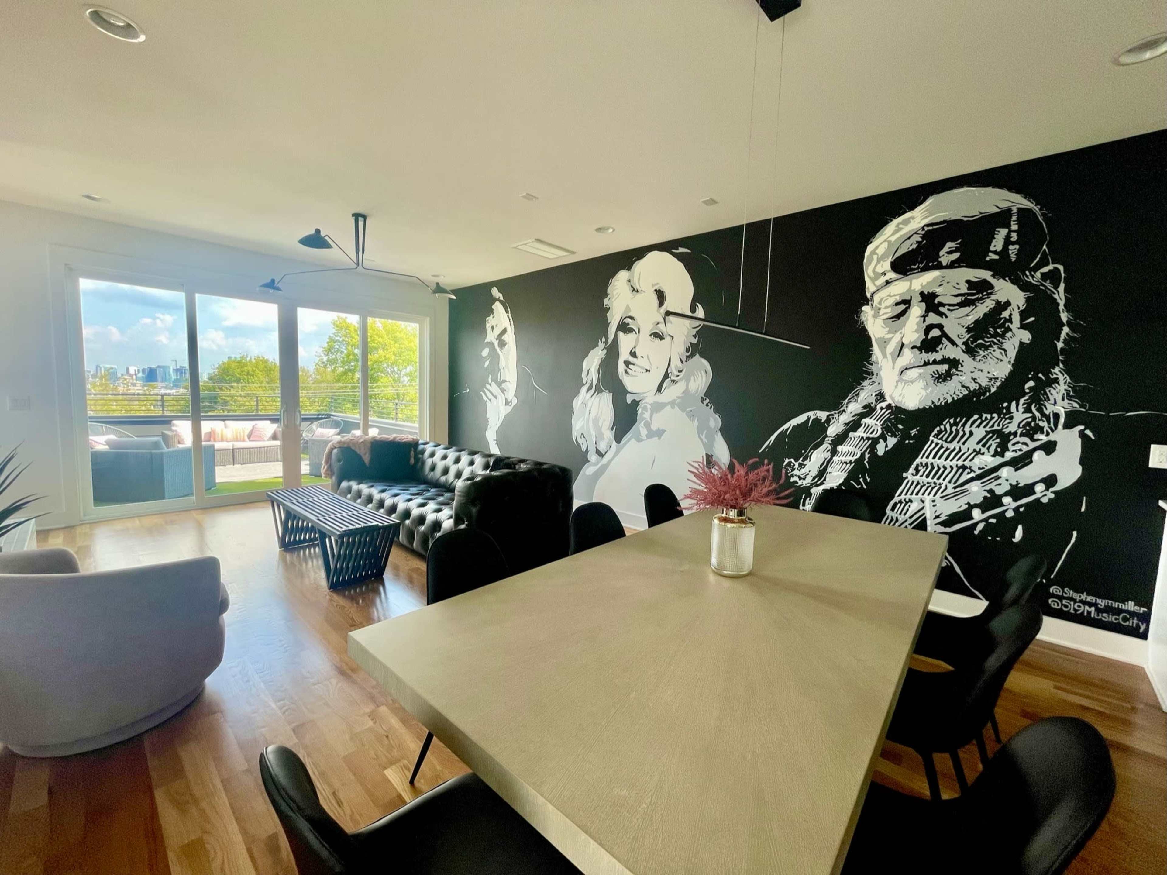 A modern living space featuring a mural of two figures on a black wall, with a dining table in the center and a view of the outdoors through large windows.