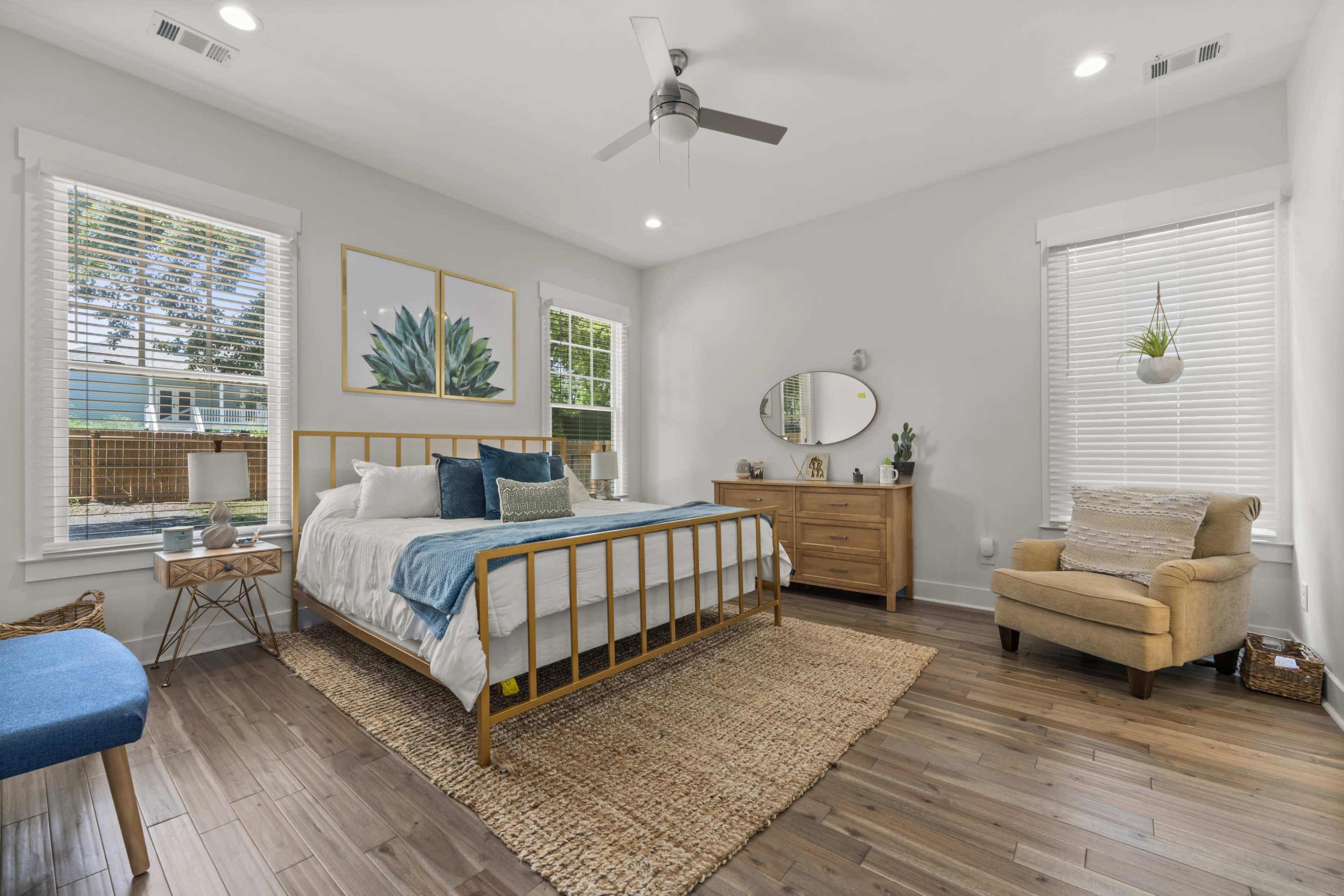 A well-lit bedroom featuring a gold metal bed frame, wooden dresser, armchair, and large windows with leafy artwork on the walls.
