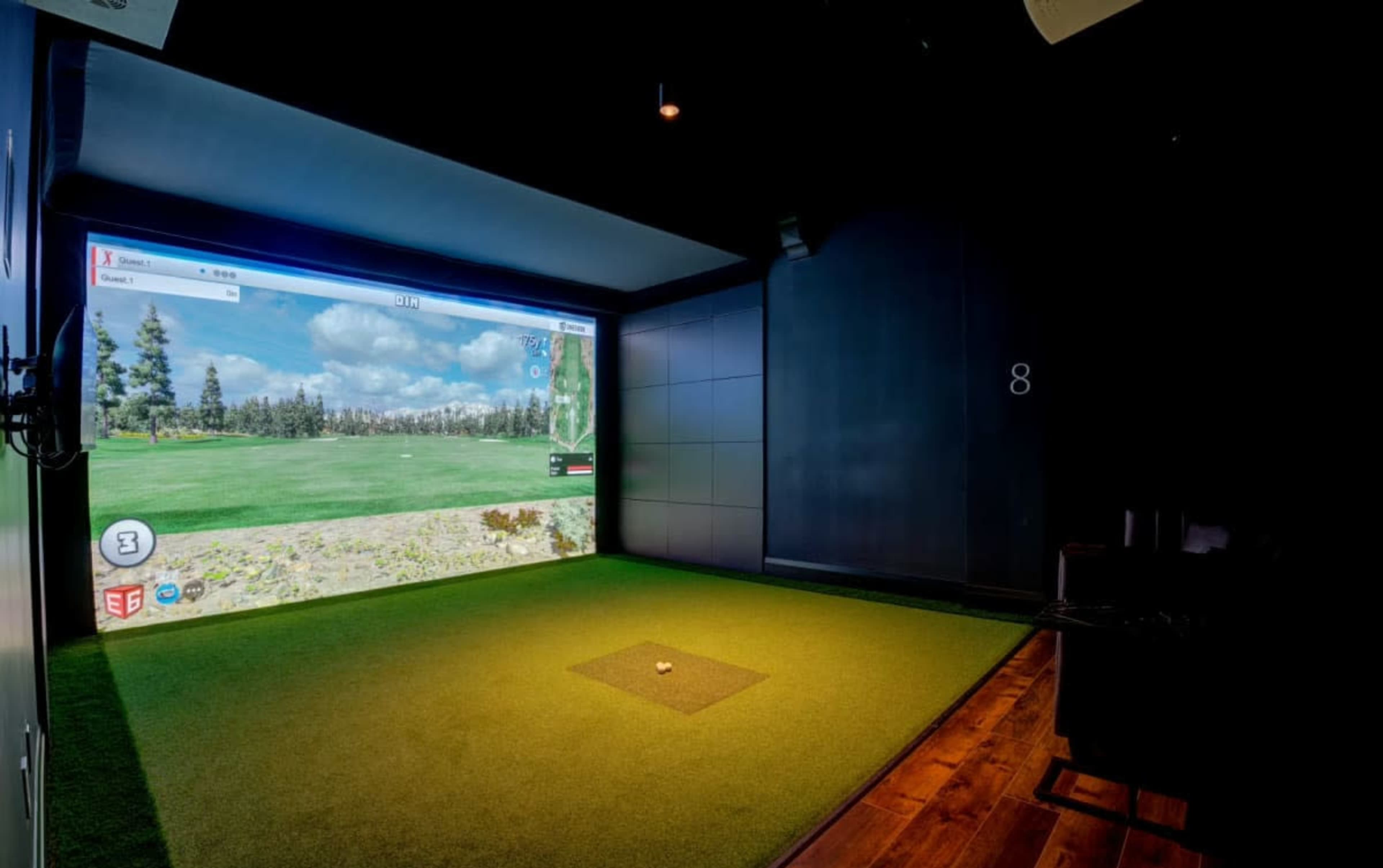 The image shows a golf simulator setup with a large projection screen displaying a golf course and artificial turf underfoot.