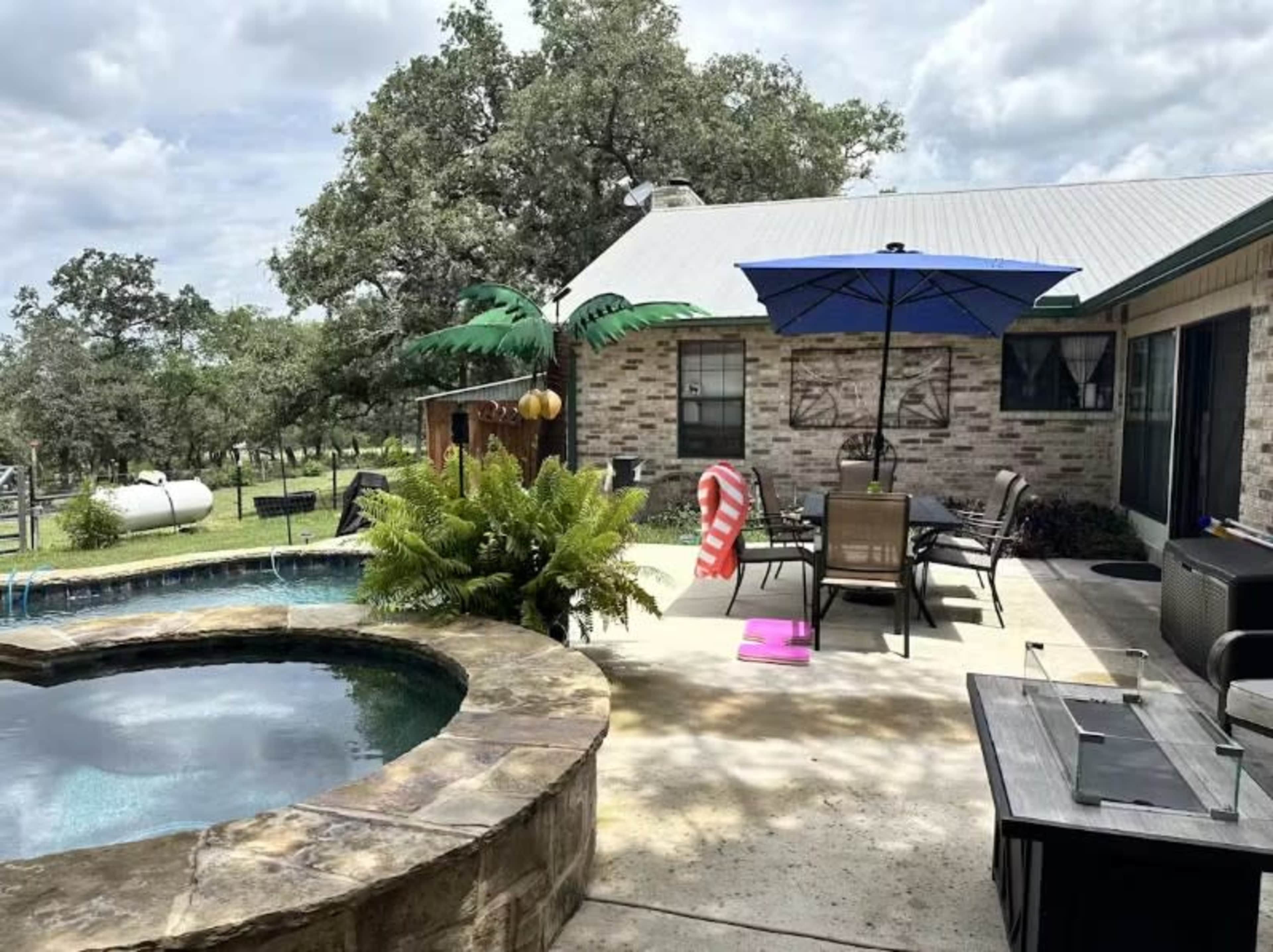 A backyard patio features a pool with a stone border, lounge chairs, an umbrella, and decorative palm trees under a partly cloudy sky.